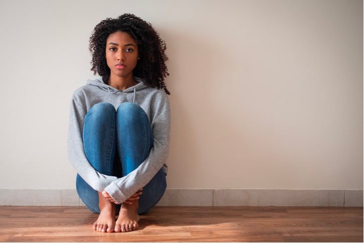 A young woman with curly hair sitting on the floor against a plain beige wall, wearing a gray hoodie and blue jeans, with her knees drawn up and her arms wrapped around them.