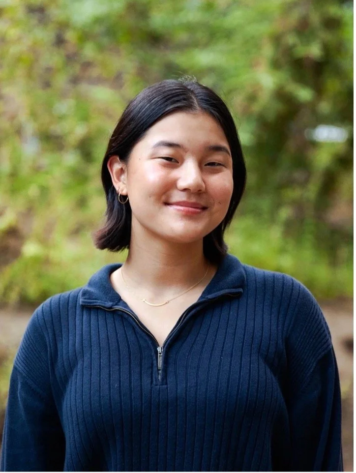 A young woman with short dark hair, wearing a navy blue zip-up sweater and small earrings, stands outdoors with a blurred green tree background, smiling gently at the camera.