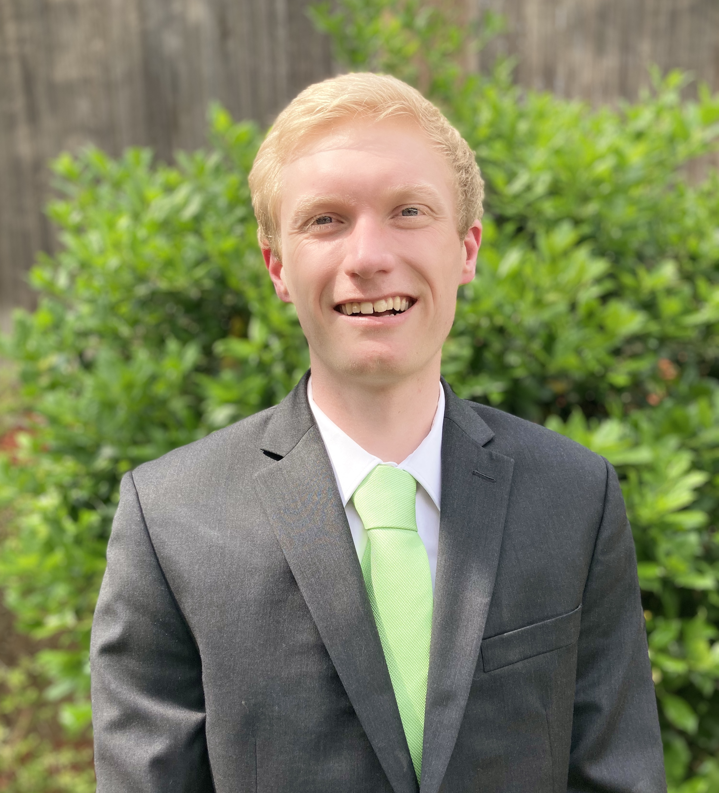 A young man with blonde hair smiling in a suit with a bright green tie, outdoor background with greenery and a wooden fence.