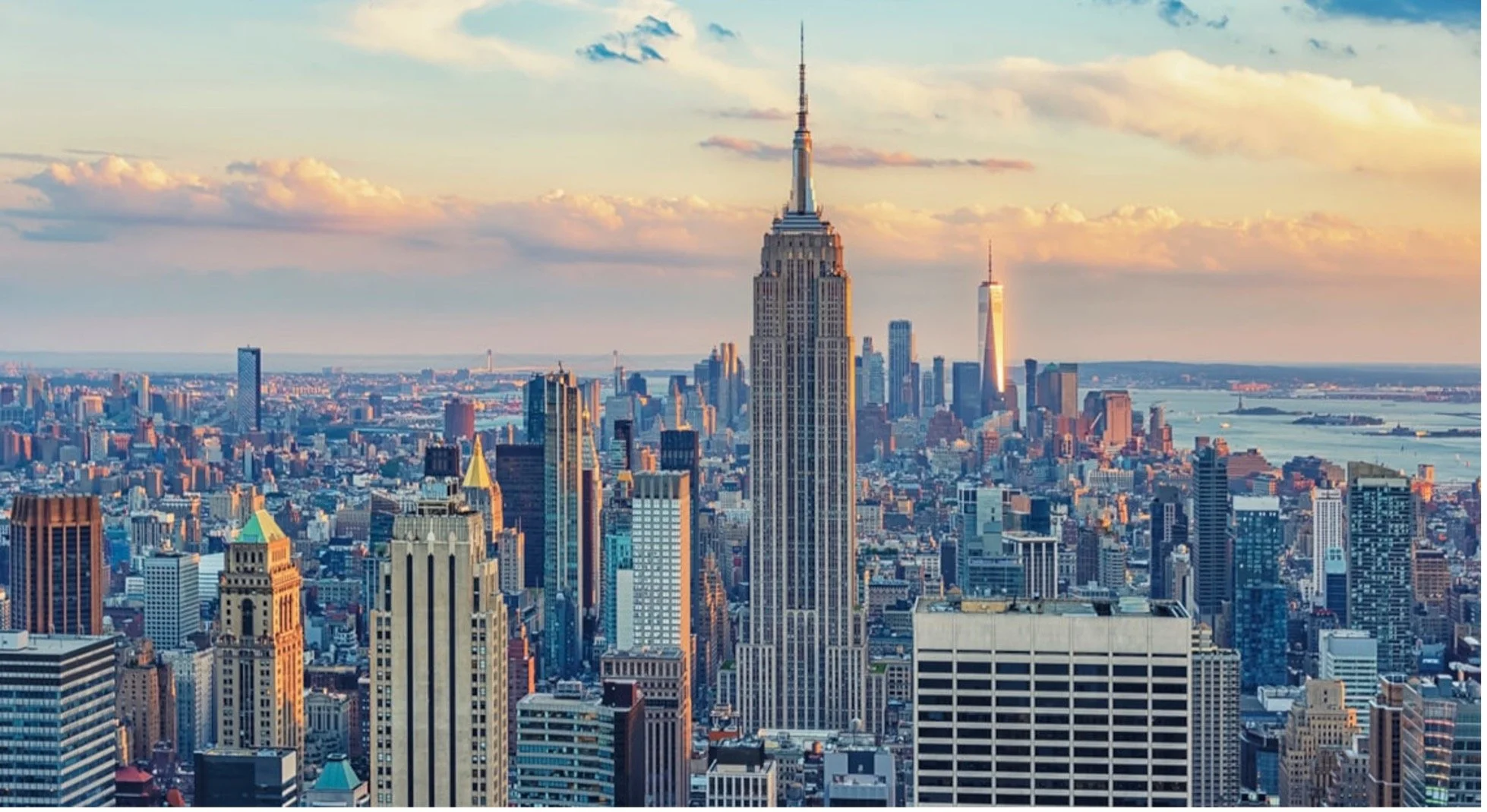 Aerial view of New York City skyline at sunset, featuring the Empire State Building and One World Trade Center among high-rise buildings with a river in the background.