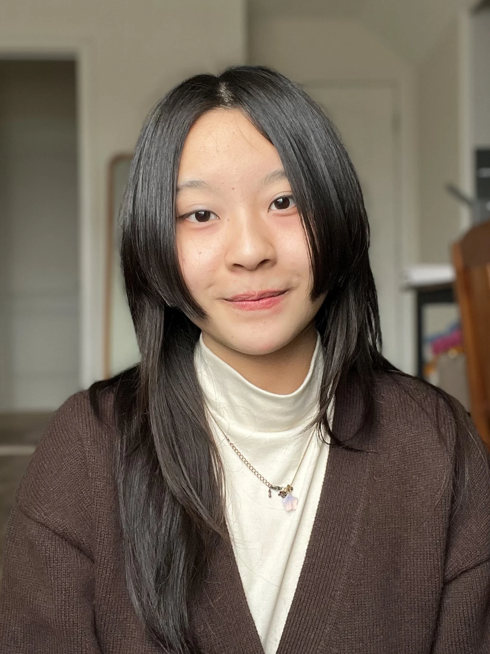 A young woman with straight dark hair, wearing a white turtleneck sweater under a brown jacket, looking at the camera with a slight smile, in a room with a blurred background including a doorway and bookshelf.