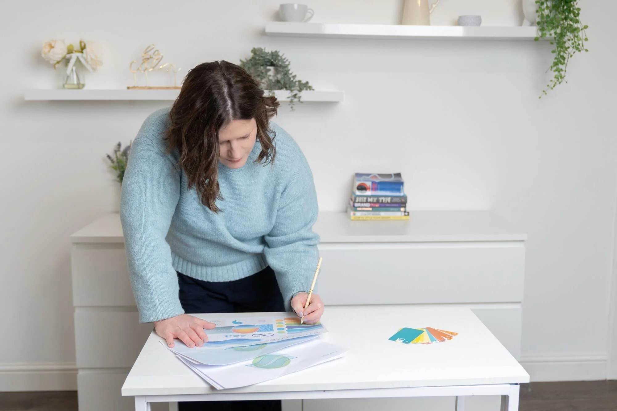 Woman working at a white desk, reviewing colour samples and charts, in a minimalistic white room with shelves, books, and plants