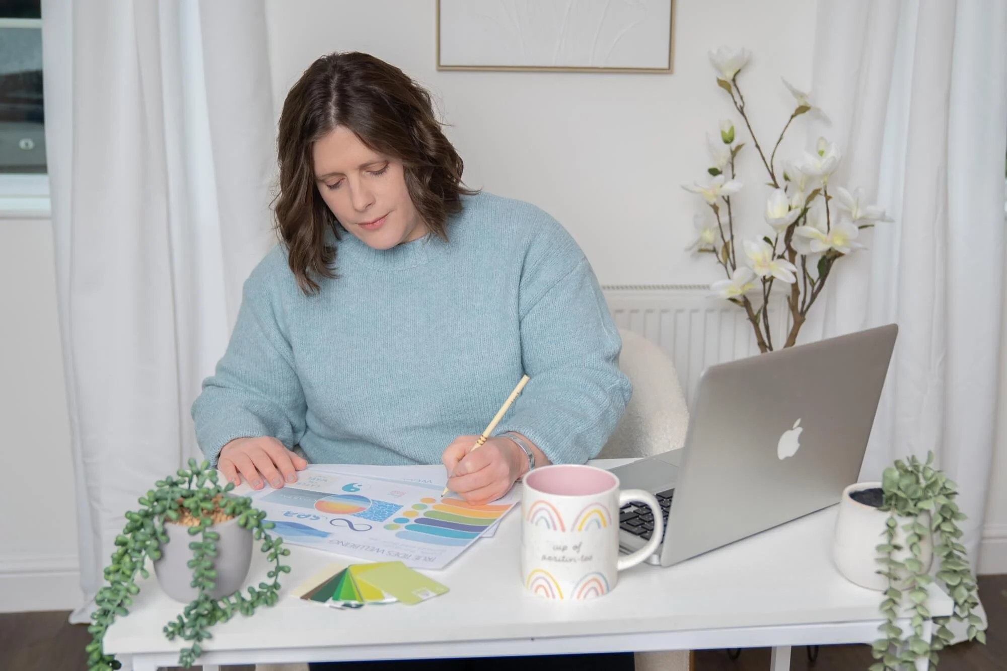 A woman with short brown hair working at a desk with plants, a coffee mug, a laptop, and colourful papers, in a bright room with white curtains and artwork.