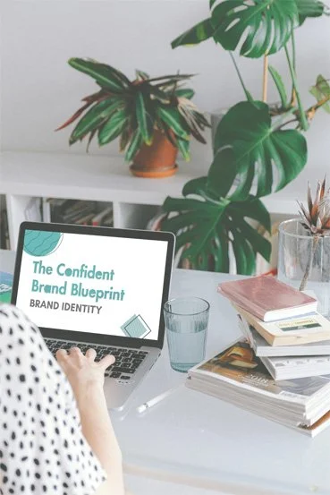 A workspace with a laptop displaying a presentation titled "The Confident Brand Blueprint." In the background, there are large leafy houseplants