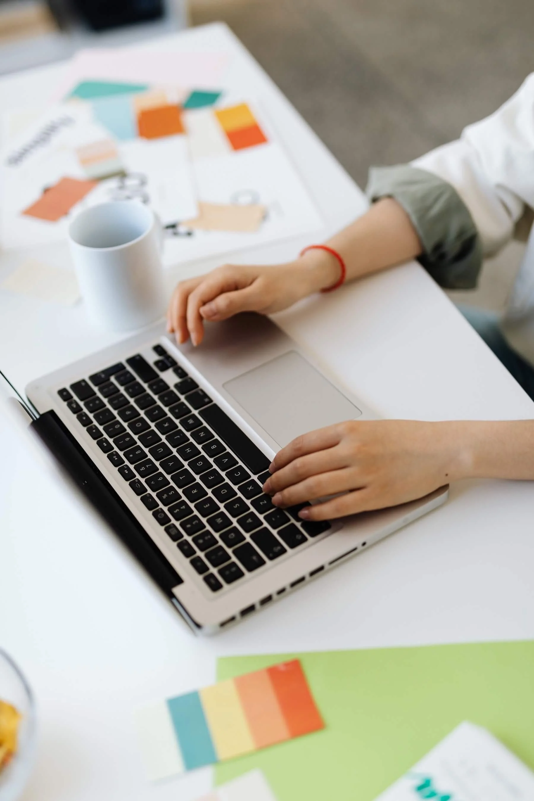 A person working at a white desk with a laptop, colourful paper swatches, a coffee mug, and paper with coloured pens