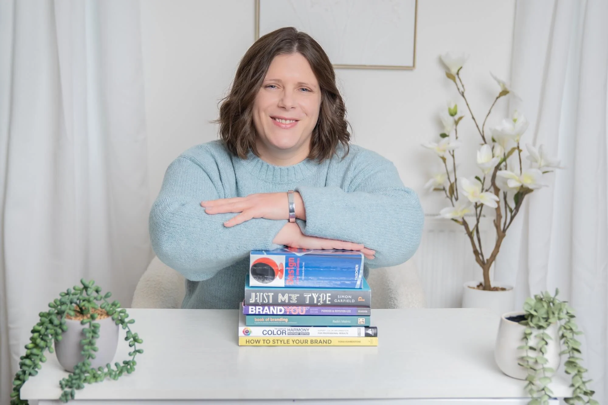 A woman with shoulder-length brown hair wearing a light blue jumper, smiling and resting her arms on a stack of books on a white table. There are plants in white pots on the table and a tall white vase with white flowers in the background