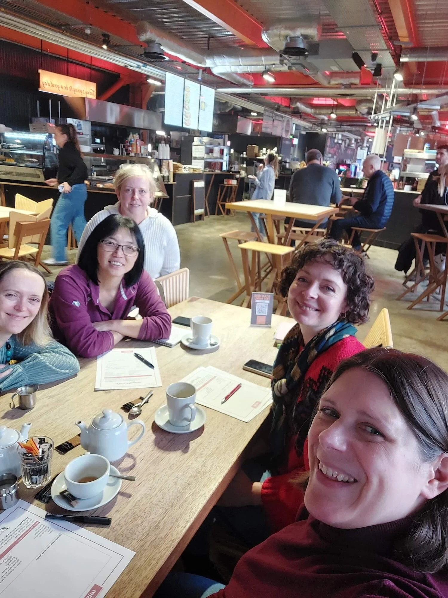 Group of people sitting at a wooden table in a cafe, smiling and posing for a photo.