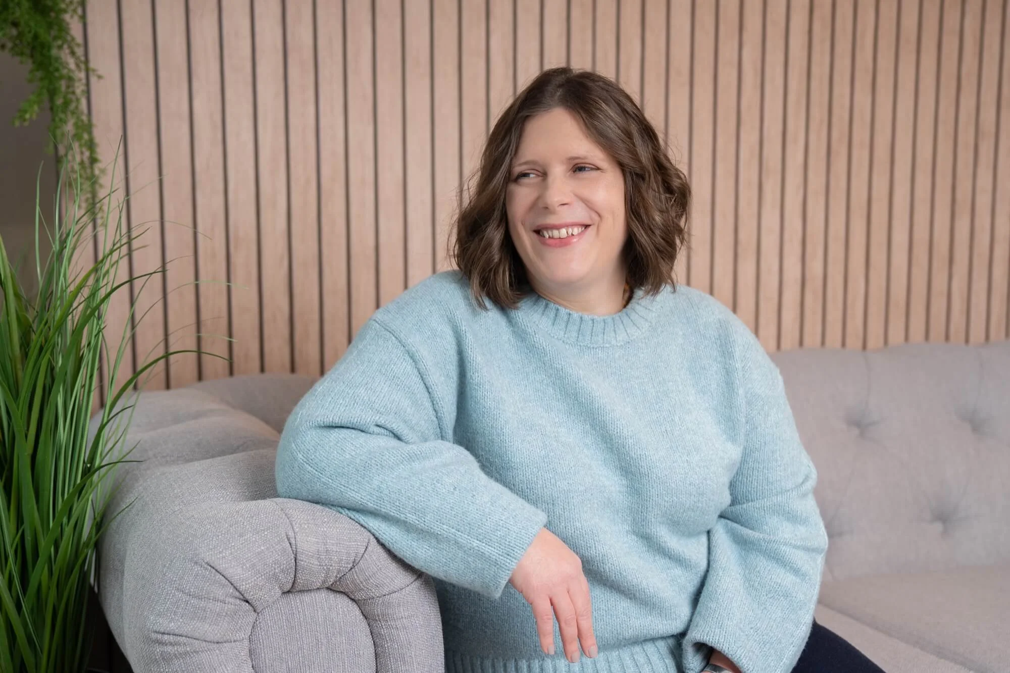 Helen Bell, a woman with shoulder-length brown hair wearing a light blue jumper, sitting on a grey sofa in a room with a wooden slat wall and a green plant to the side