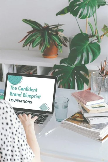 A person working on a laptop with a screen that reads 'The Confident Brand Blueprint Foundations,' surrounded by stacks of books, a glass of water, and houseplants in a bright, organised workspace