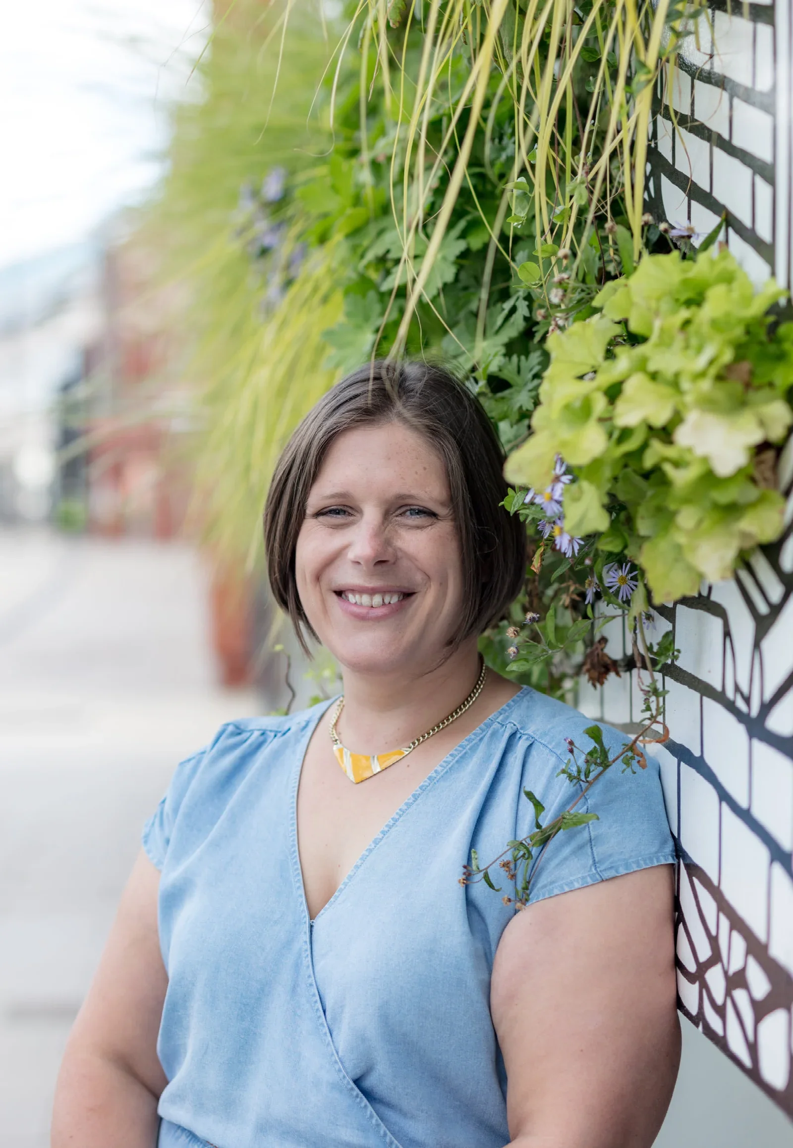 A woman with short brown hair smiling, wearing a light blue top, standing outdoors beside a wall covered with foliage