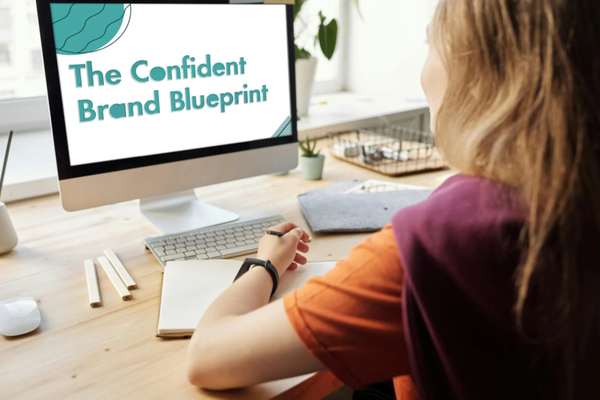 A woman sitting at a desk looking at a computer screen displaying the text 'The Confident Brand Blueprint'