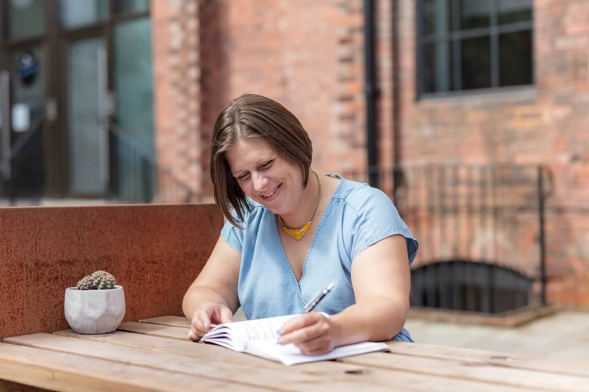 Helen Bell, a woman with short brown hair, smiles while writing in a notebook at an outdoor wooden table, with a brick building in the background