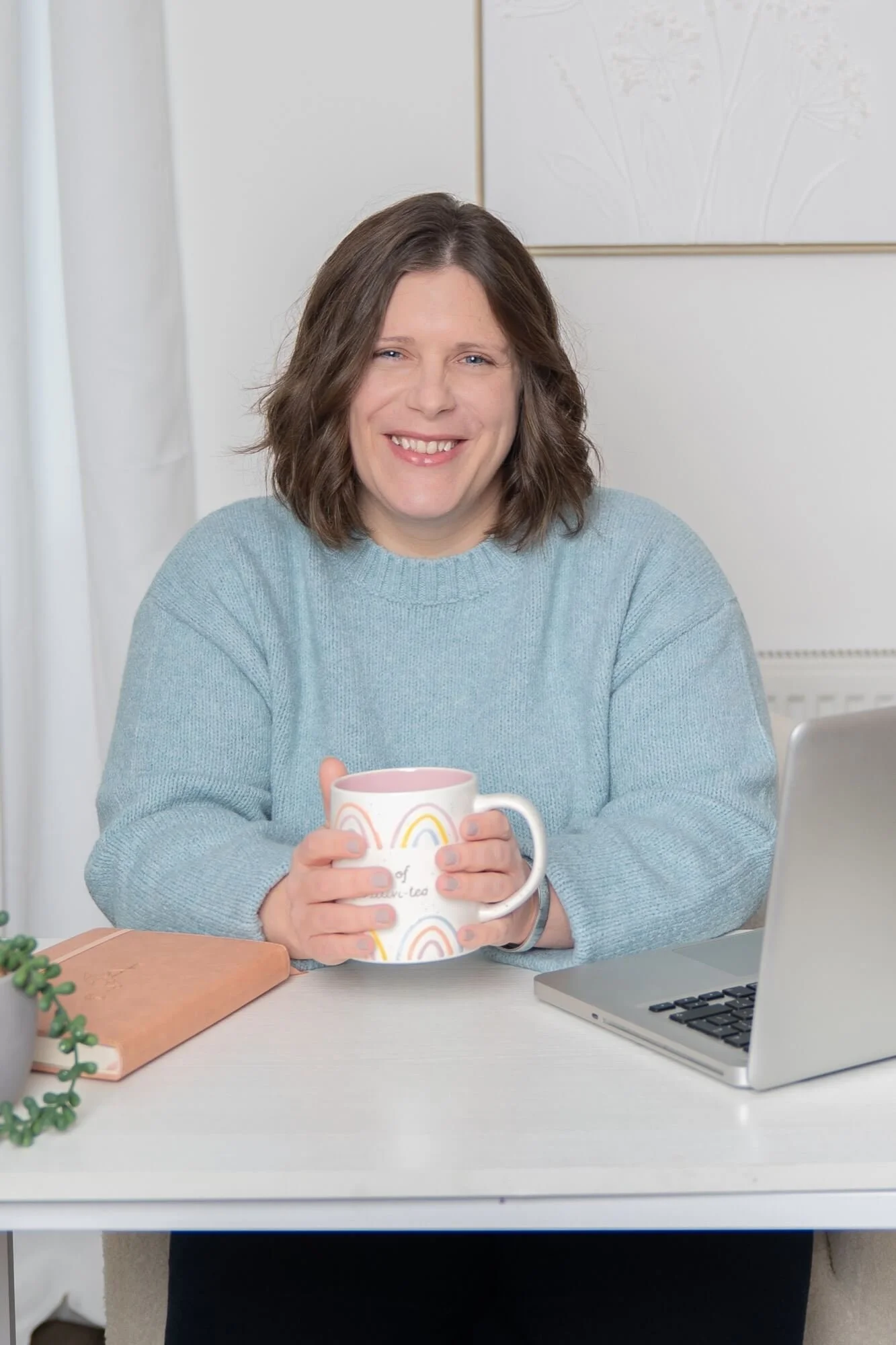 A woman with shoulder-length brown hair wearing a light blue jumper, sitting at a white desk and holding a mug, smiling