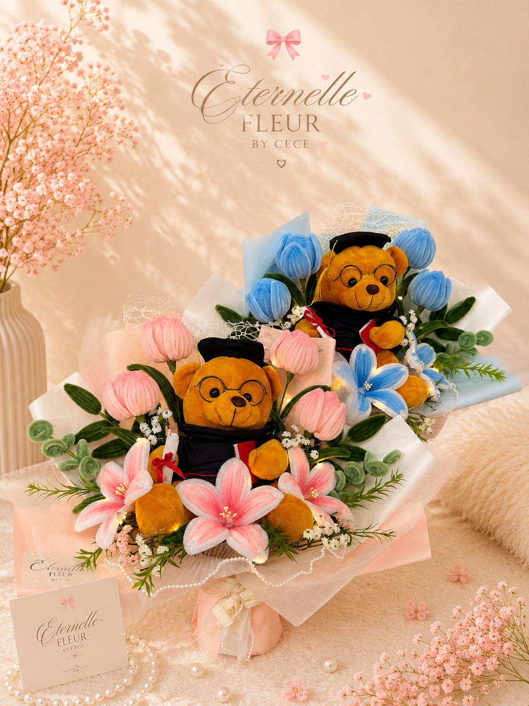 A floral arrangement with two plush teddy bears dressed in graduation caps and gowns, surrounded by pink and blue flowers, placed on a beige table with pink flowers and pearl accessories, and a pink vase with pink blossoms in the background.