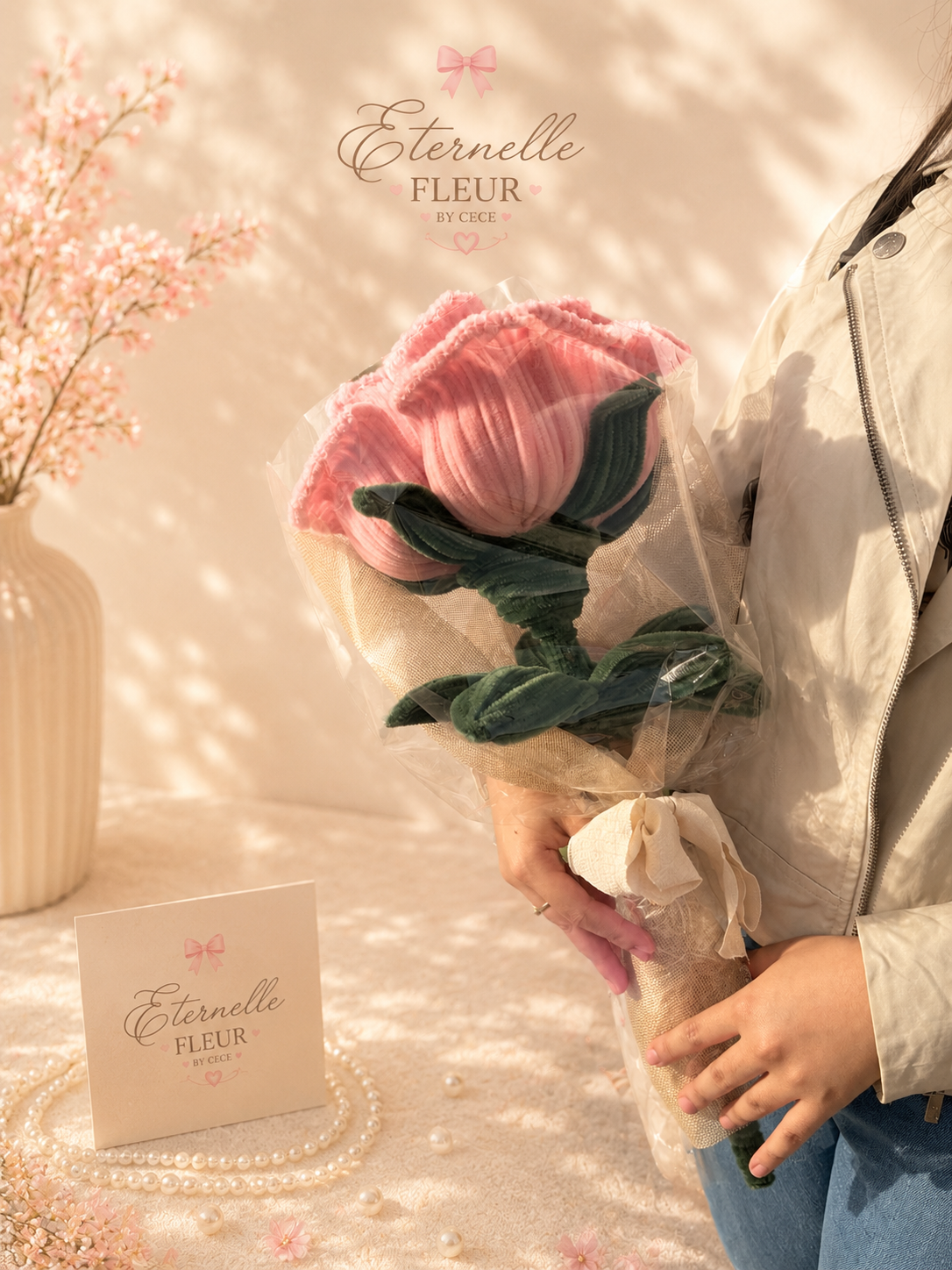 Person holding a bouquet of pink and green fabric flowers wrapped in clear plastic, with a pink ribbon tying the wrapping. A card with the text 'Eternelle Fleur by Cece' is on a table decorated with pearls and pink flower petals, with a vase of pink 