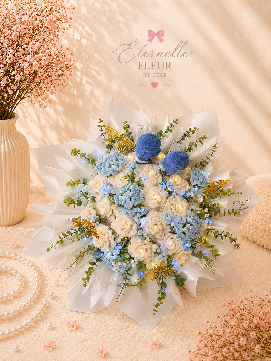 A bouquet of white roses and blue flowers decorated with green leaves, small white lights and two large blue pom-poms, set on a soft pink surface surrounded by pink flowers, a pearl necklace, and a beige pillow. A beige vase with pink flowers is in the background, with a backdrop bearing the words 'Eternelle Fleur by Cece' and a pink bow.