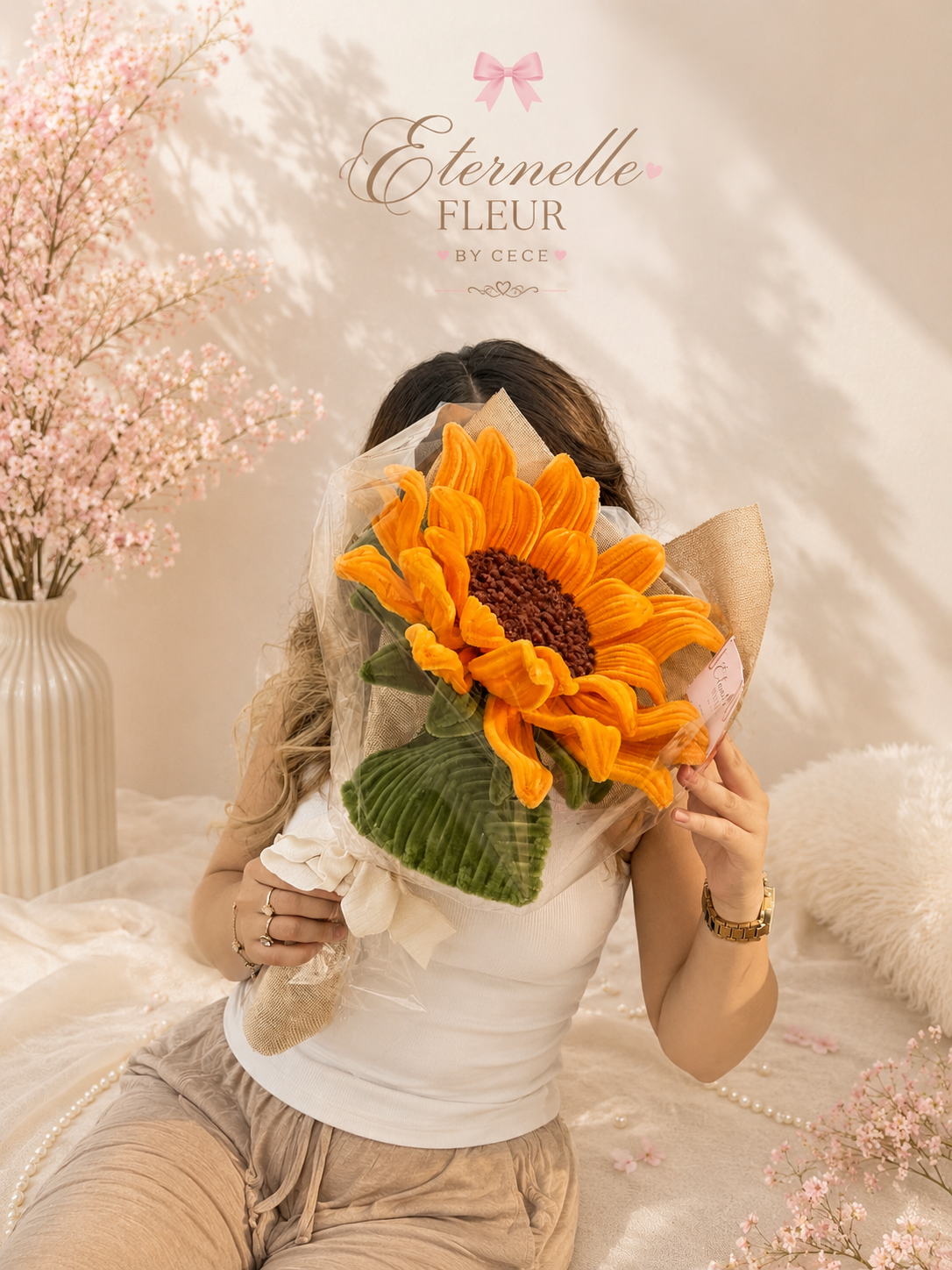 Woman holding a large sunflower bouquet in front of her face, sitting on bed with pink flowers to the side, soft beige background, and the text 'Eternelle Fleur by Cece' at the top.
