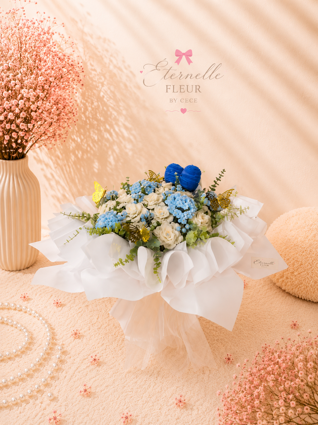 A pink and white floral arrangement featuring blue and white flowers with green foliage, wrapped in white tissue paper. The display is on a peach-colored surface, surrounded by pink baby's breath flowers, a cream-colored striped vase, pearl necklaces, and a peach plush cushion. A pink bow and the words 'Eternelle Fleur by Cece' are on the pale pink background.