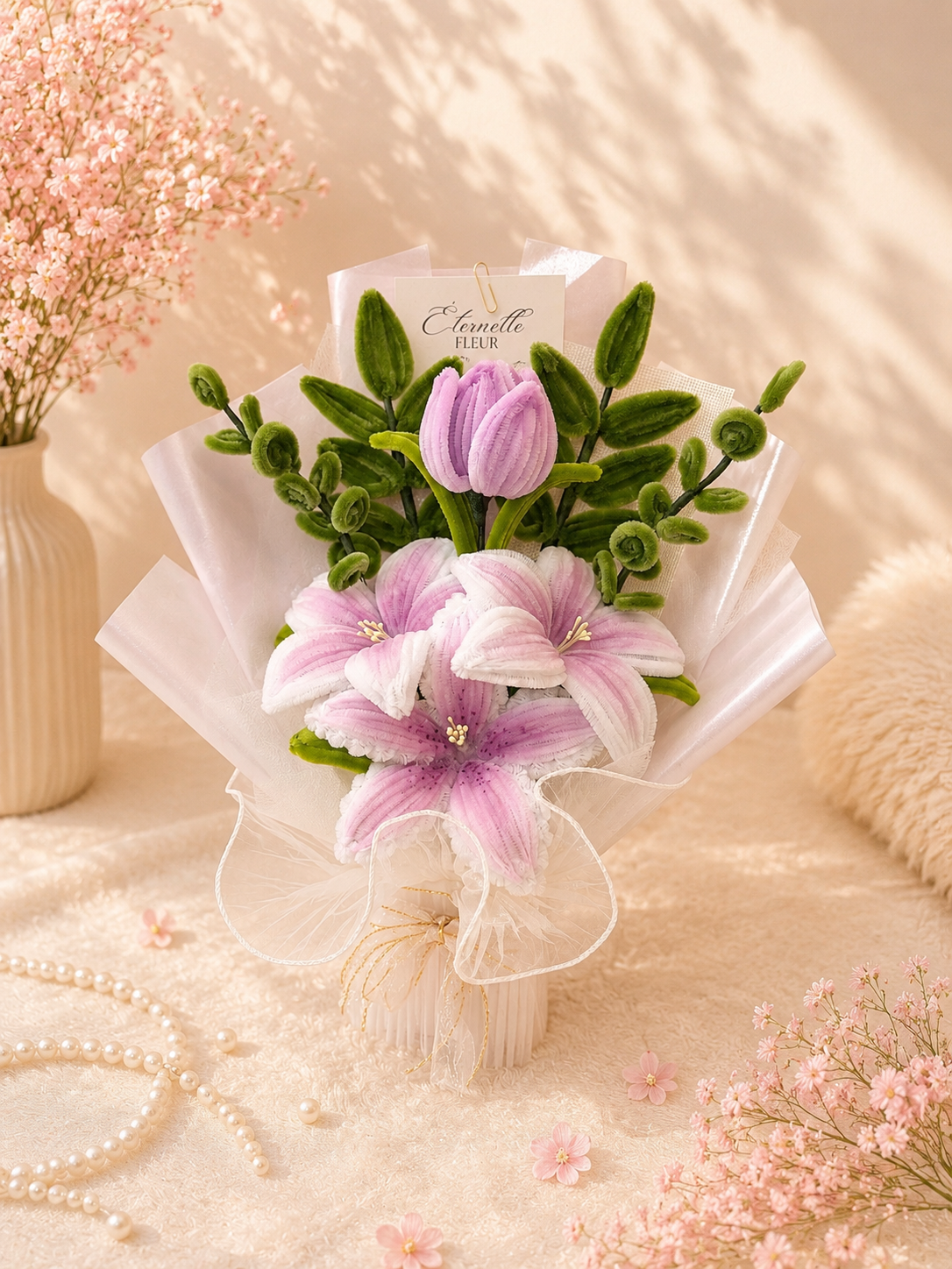 Pink and white artificial flower bouquet with lilies and tulips in a cream vase, surrounded by pink flowers, pearls, and soft fabric.