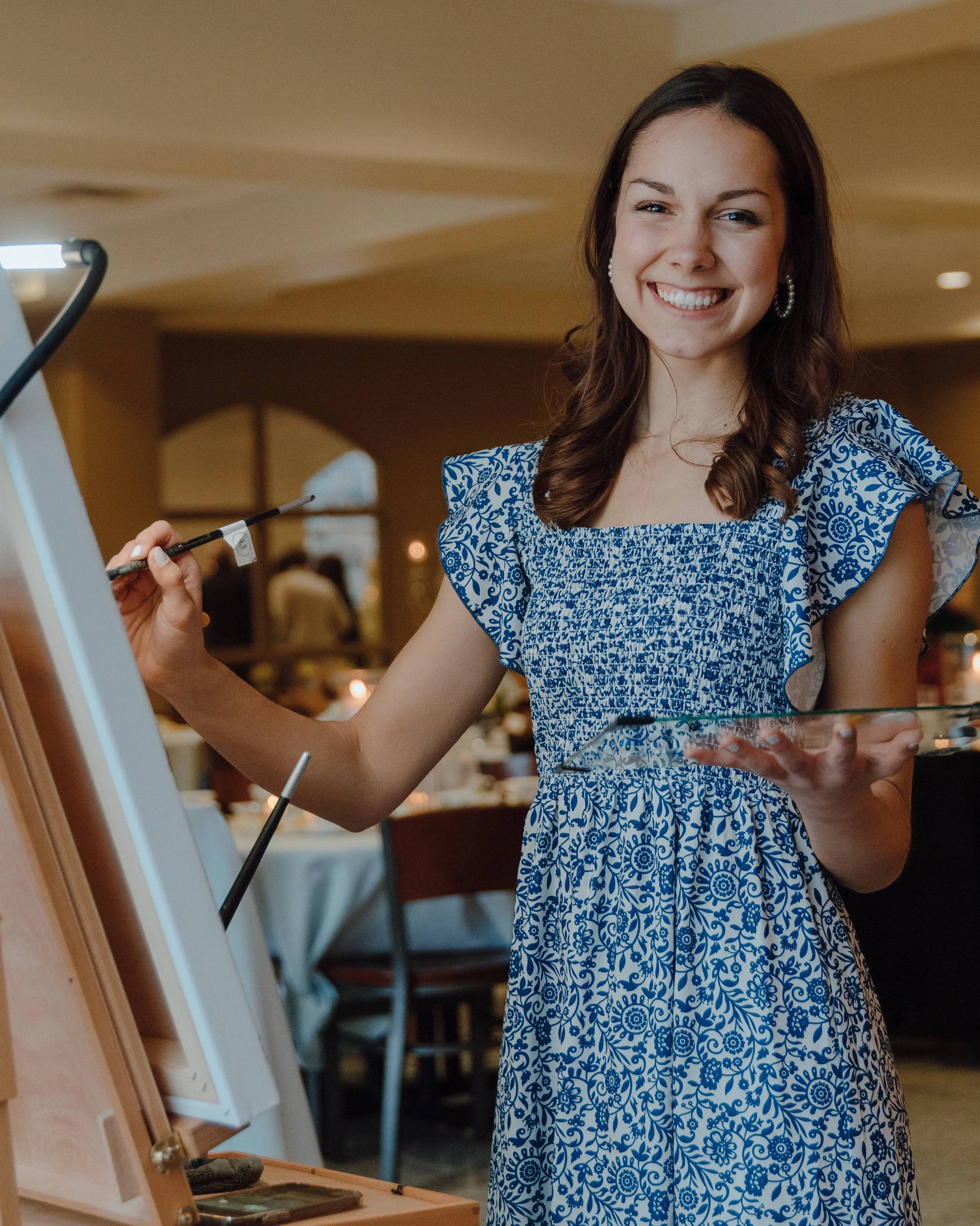 A smiling woman in a blue and white patterned dress standing in a room, possibly at a social event or gathering.
