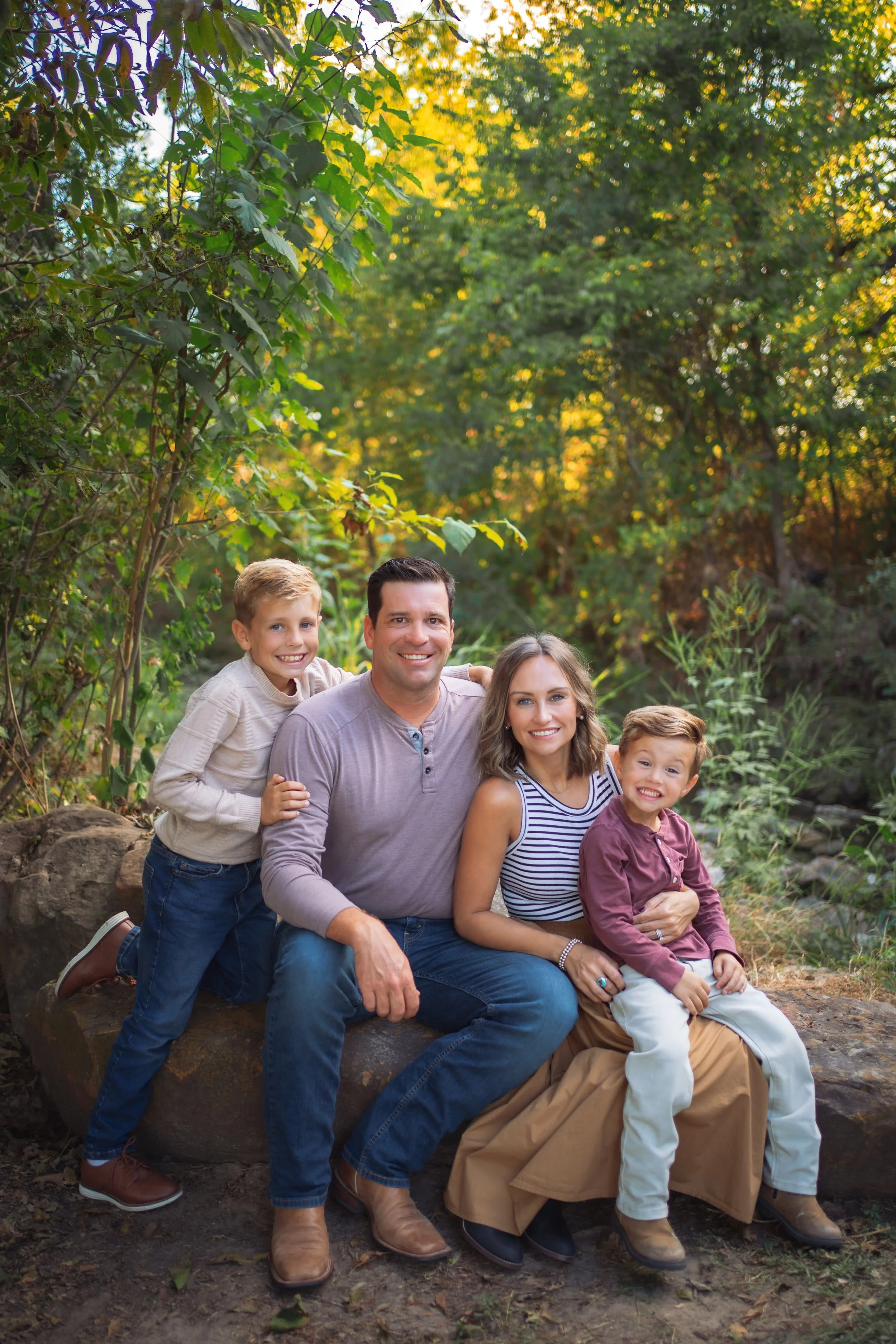 A family of five sitting outdoors in a natural setting with trees and greenery, smiling for a group photo.