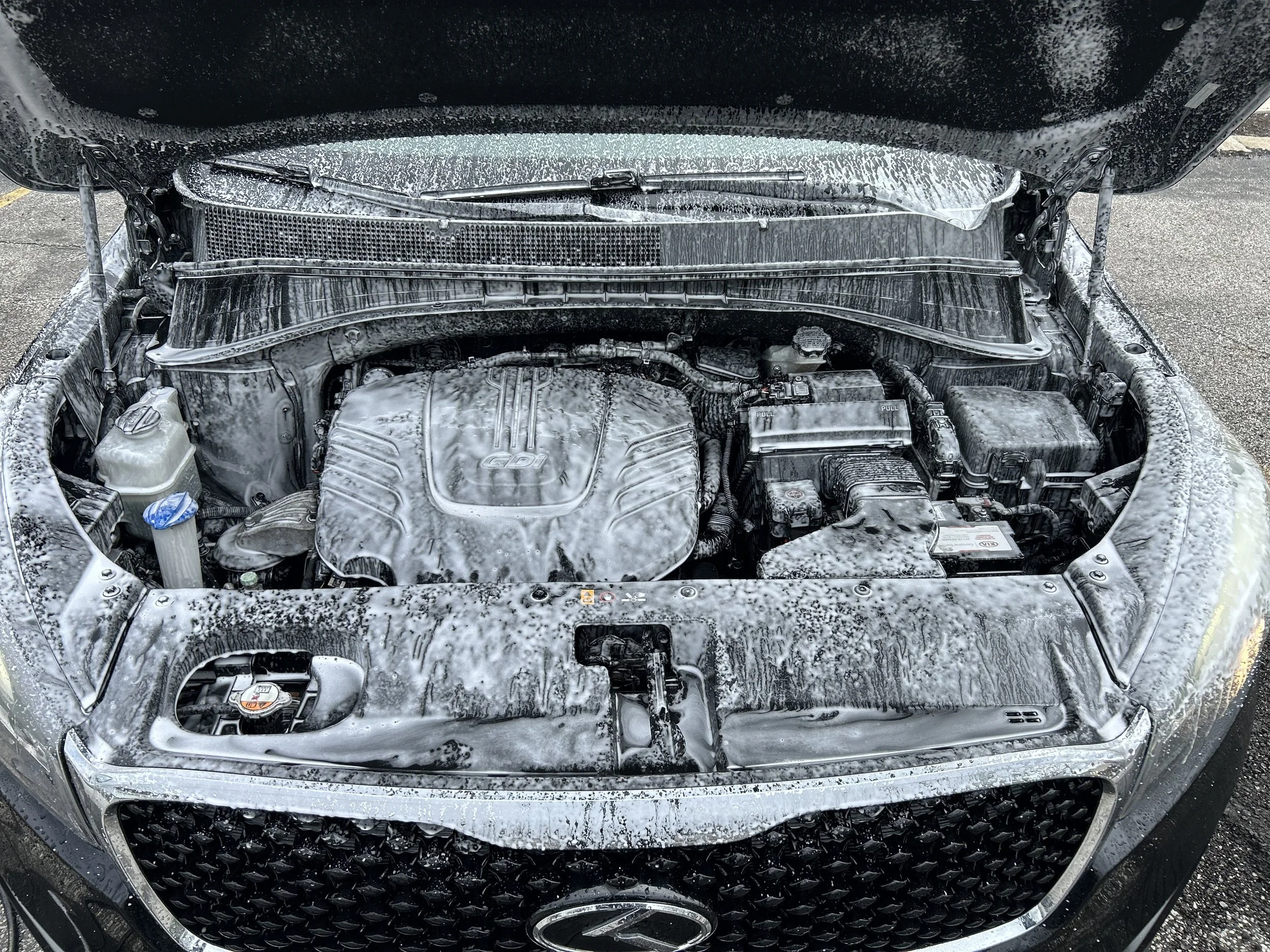 Car engine bay covered in soap suds during washing, with foam dripping down the engine components and hood.