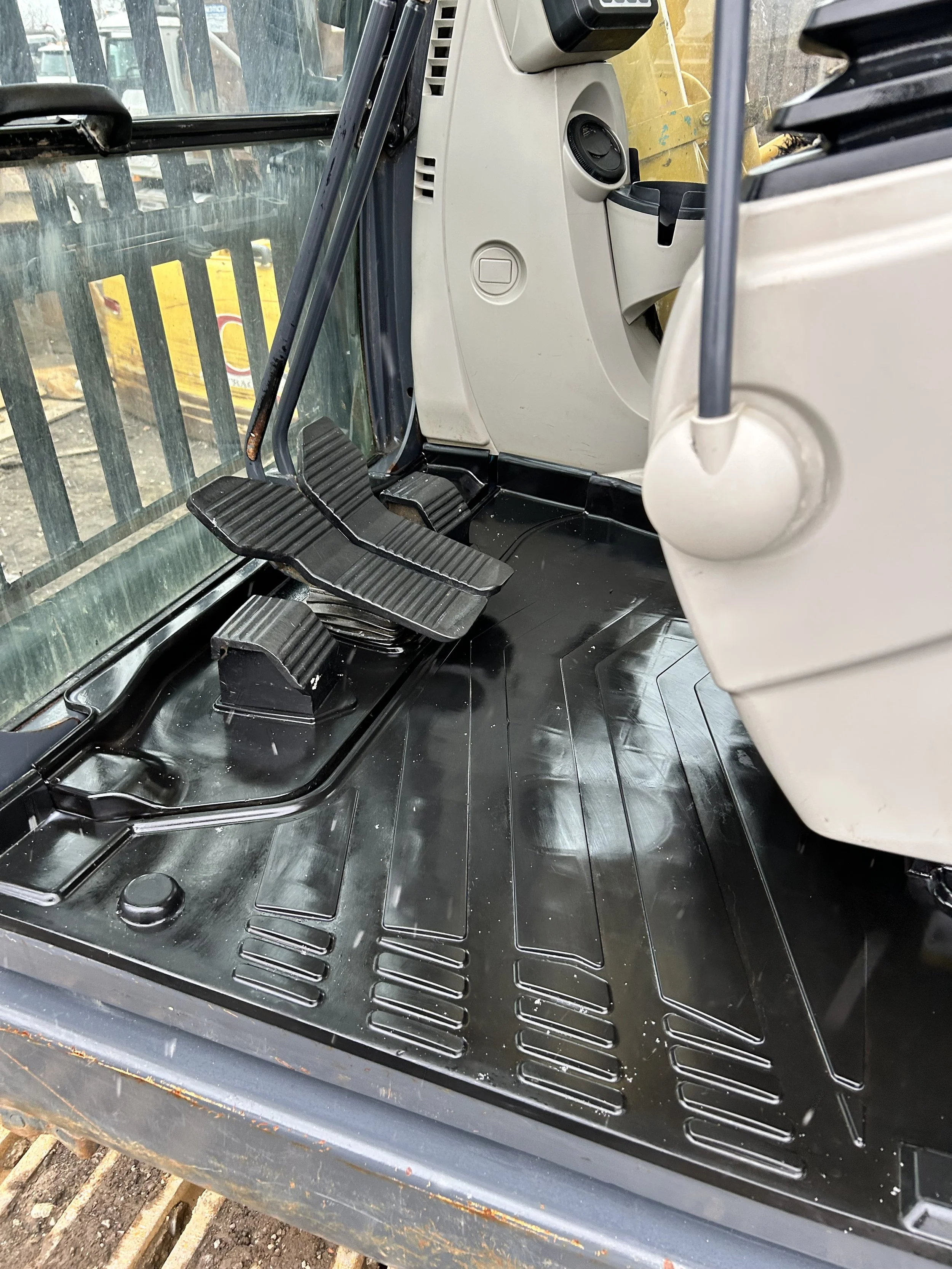 Close-up of the interior of a construction vehicle, showing black foot pedals, a beige dashboard, and large windows with a construction site visible outside.