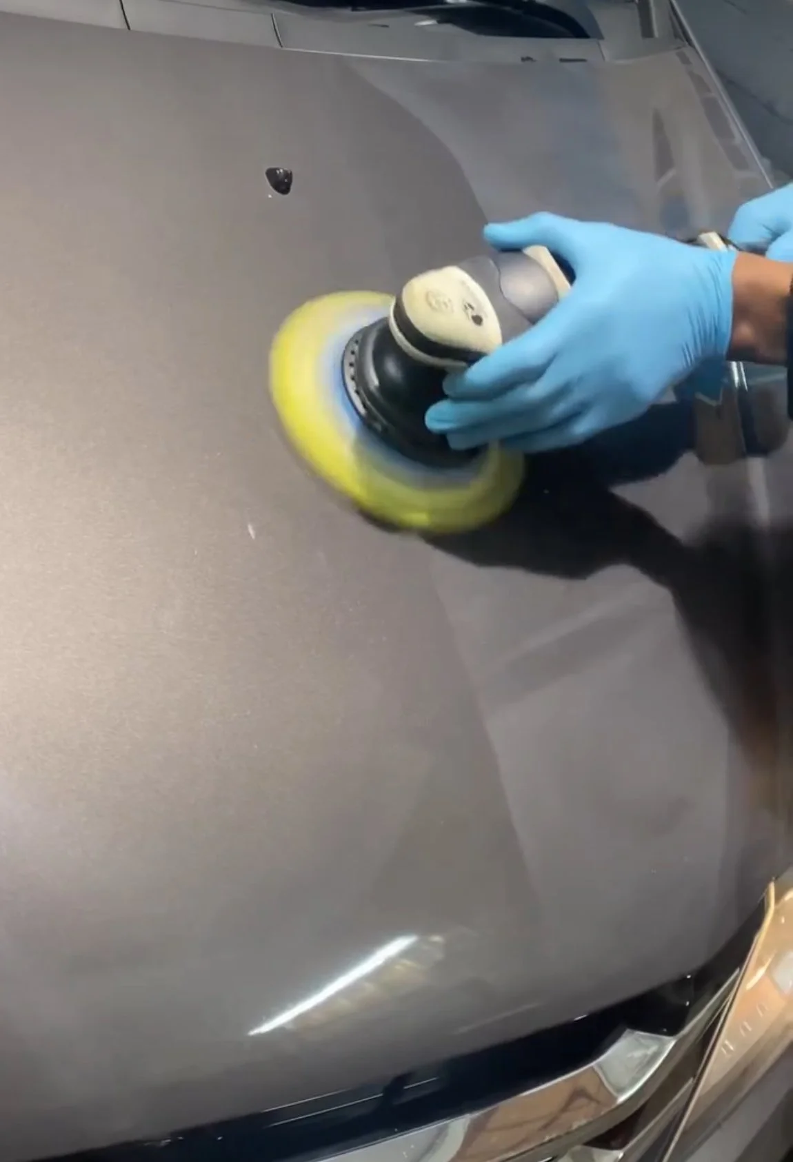 Person wearing blue gloves using a power buffer to polish a gray car's surface.