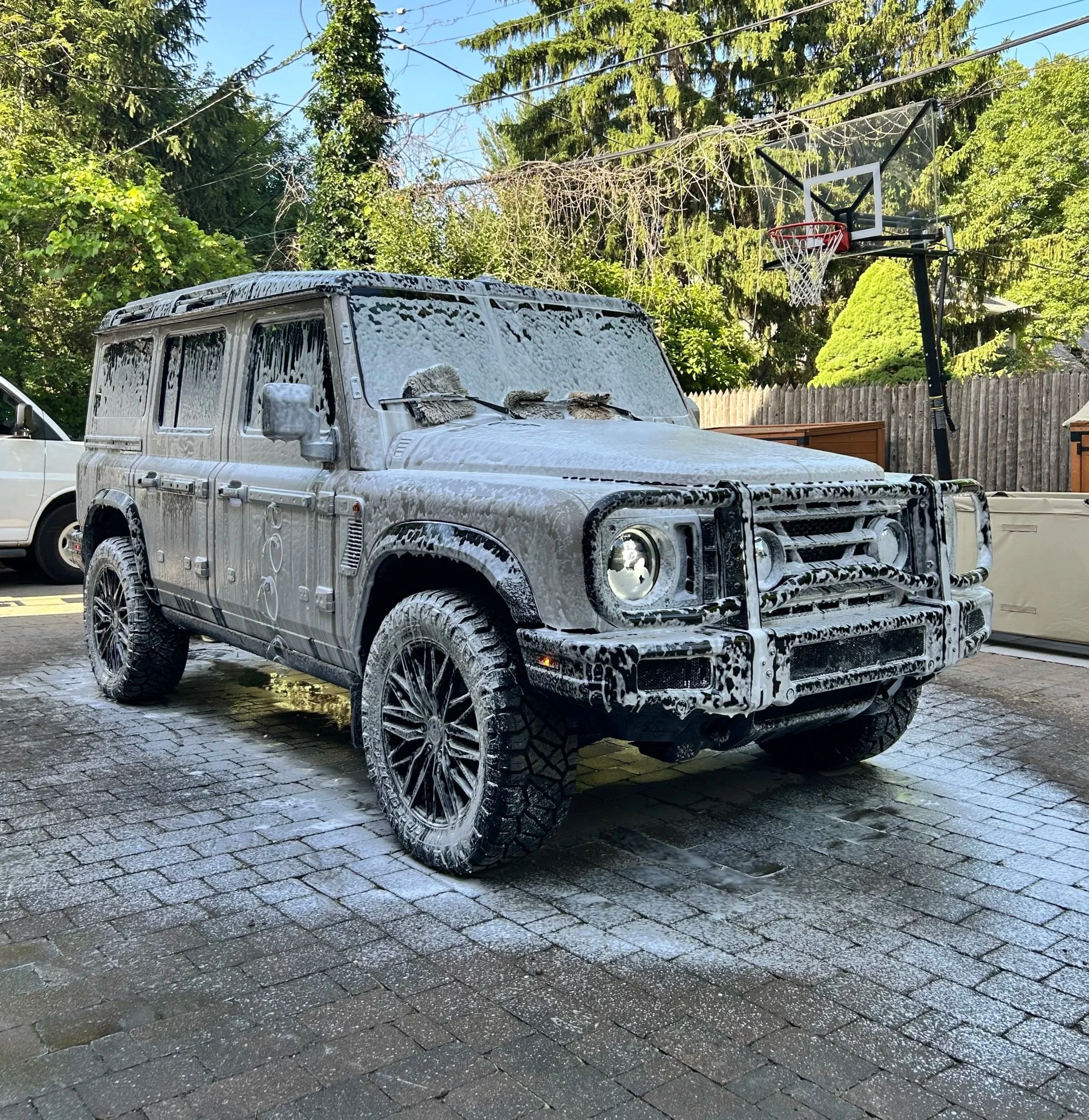 A white SUV covered in soap suds being washed, parked on wet pavement outside with trees and a basketball hoop in the background.