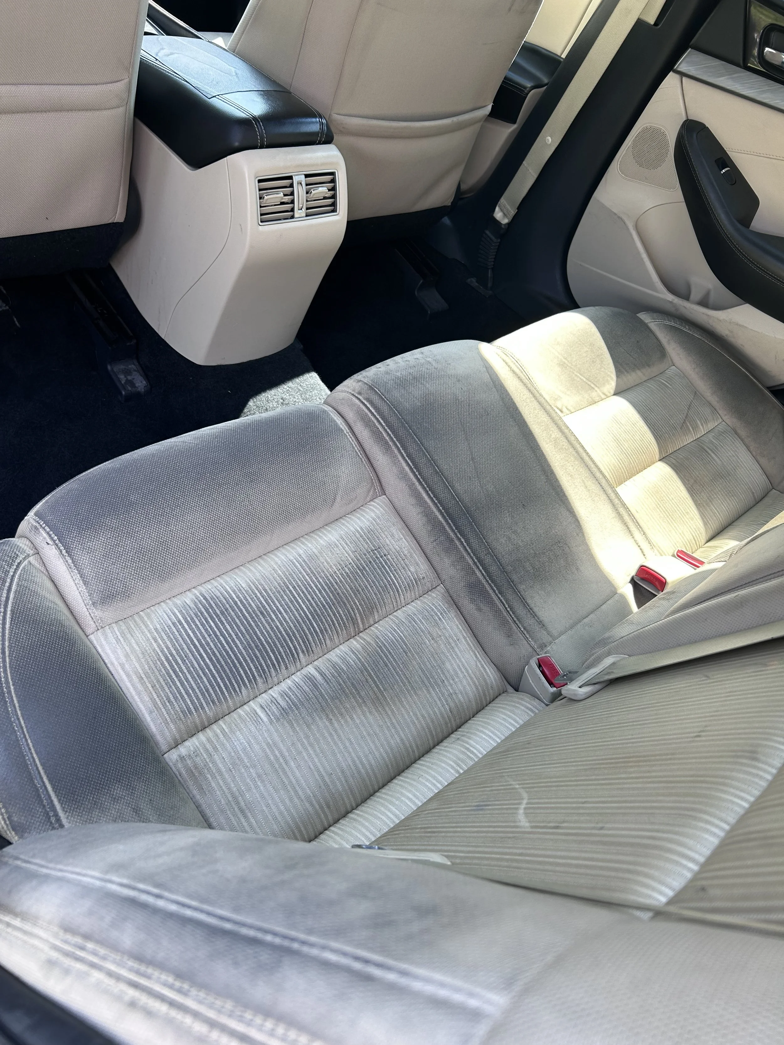 Rear interior of a car showing the backseat with fabric upholstery, seat belts, and the back of the front seats with air vents.