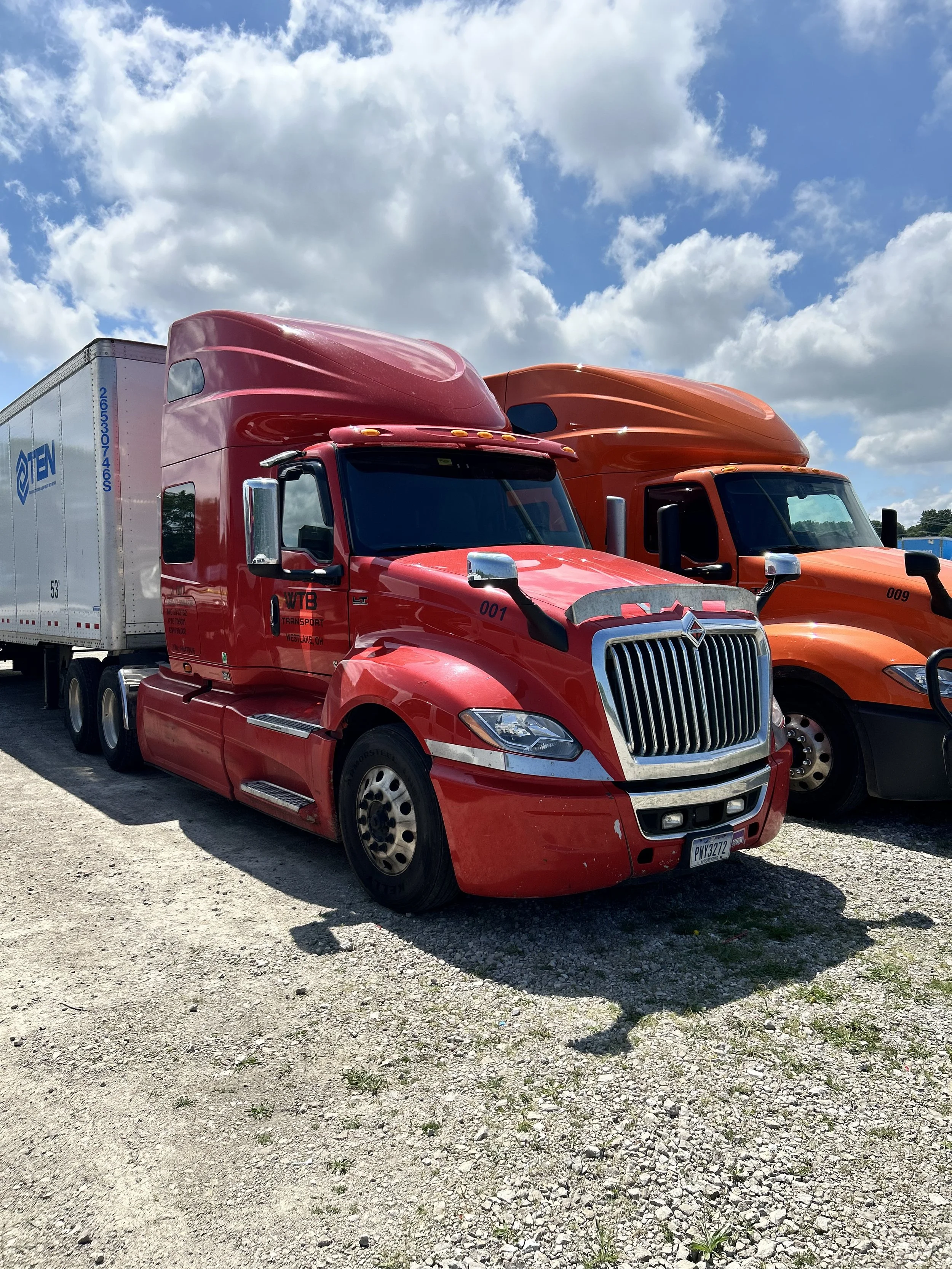 A row of semi-trailer trucks parked on a gravel lot under a partly cloudy sky, with a red truck in the foreground and orange trucks behind.