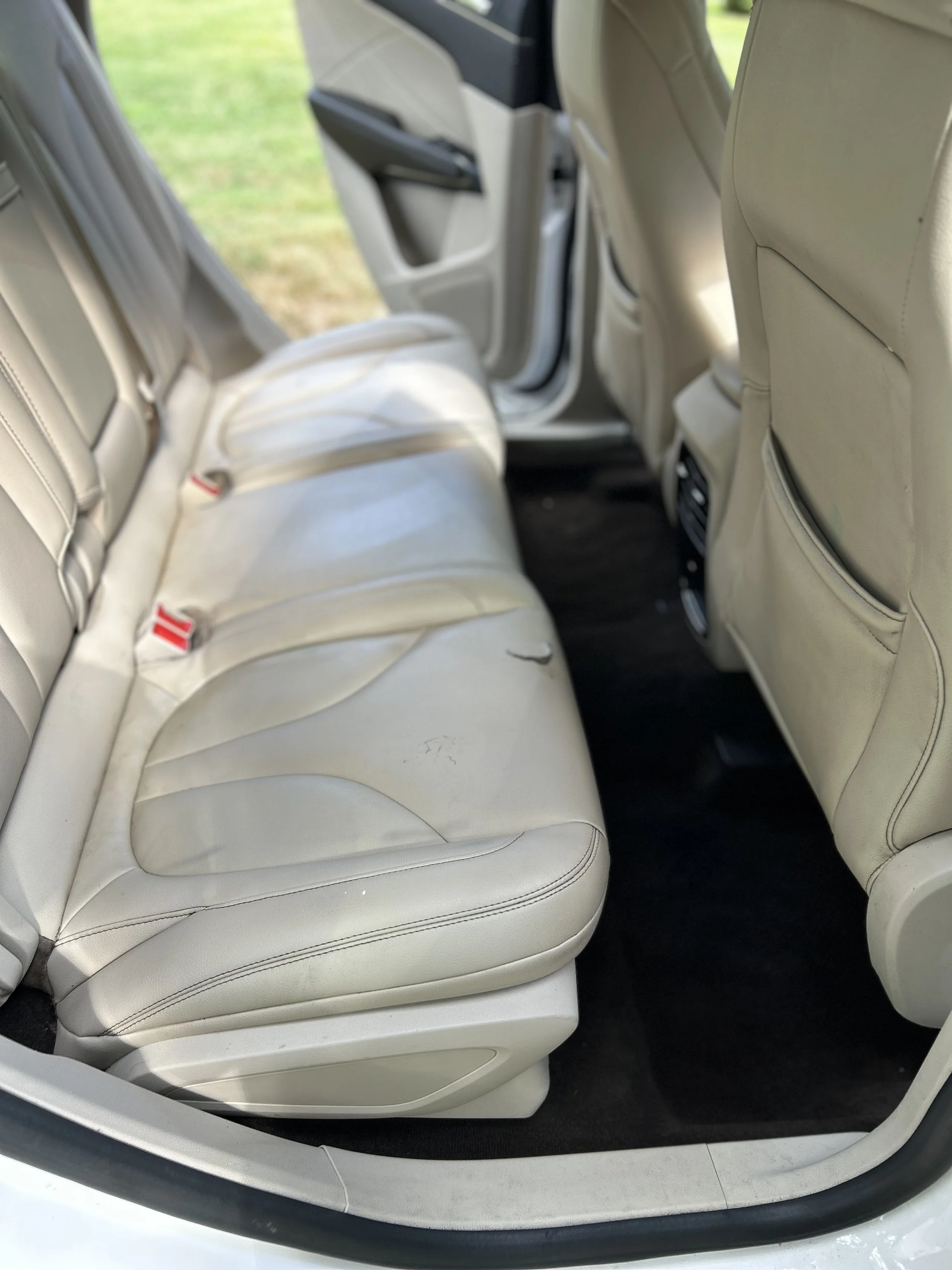 The interior of a vehicle showing cream-colored leather back seats and the rear of the front passenger seat, with the rear door open and grass outside visible.