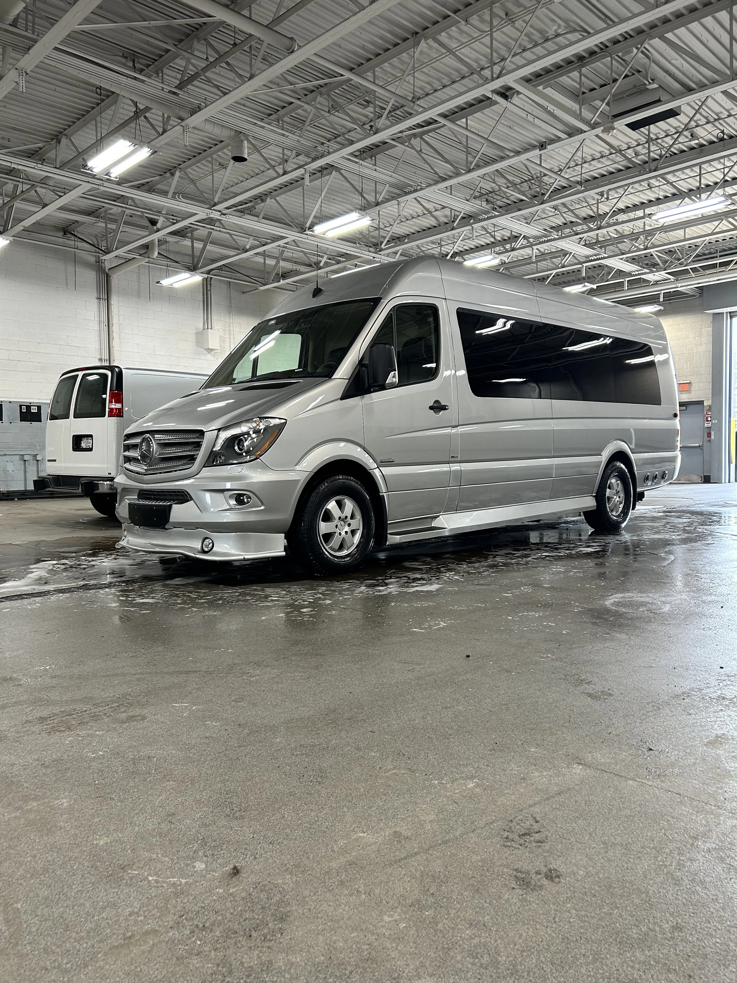 A silver passenger van parked inside a large industrial garage or warehouse with a high ceiling and fluorescent lighting.