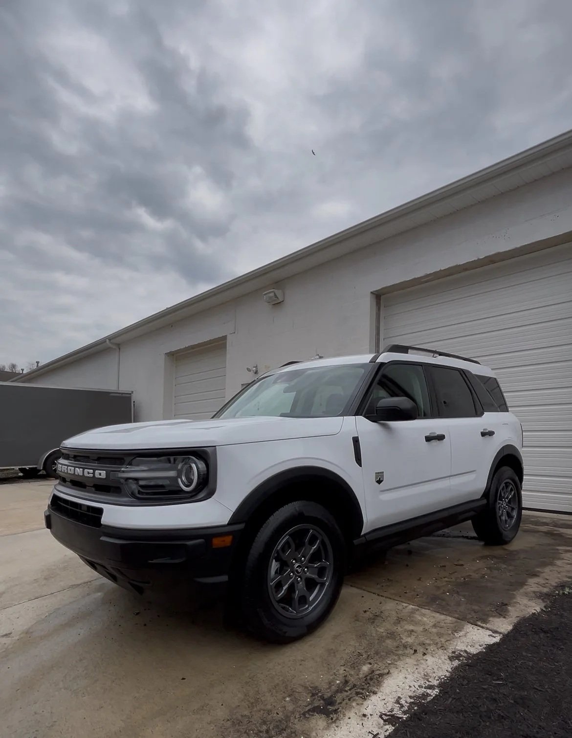 White Ford Bronco SUV parked outside a building with a white exterior and garage doors, under a cloudy sky.