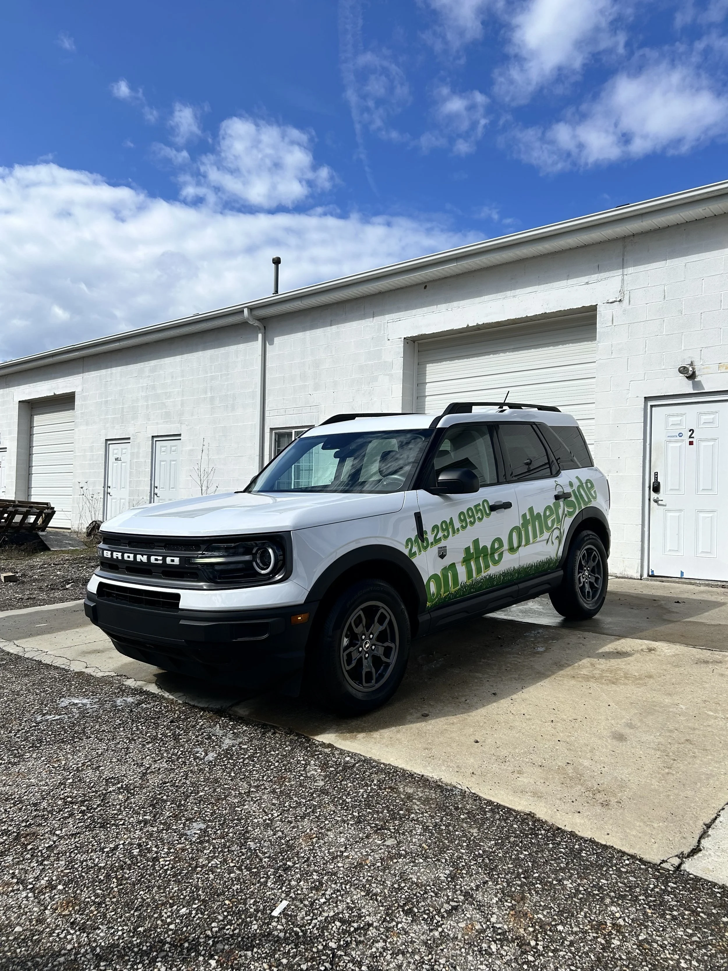 A white Ford Bronco parked outside a white industrial building with closed garage doors. The sky is partly cloudy with a bright blue background.