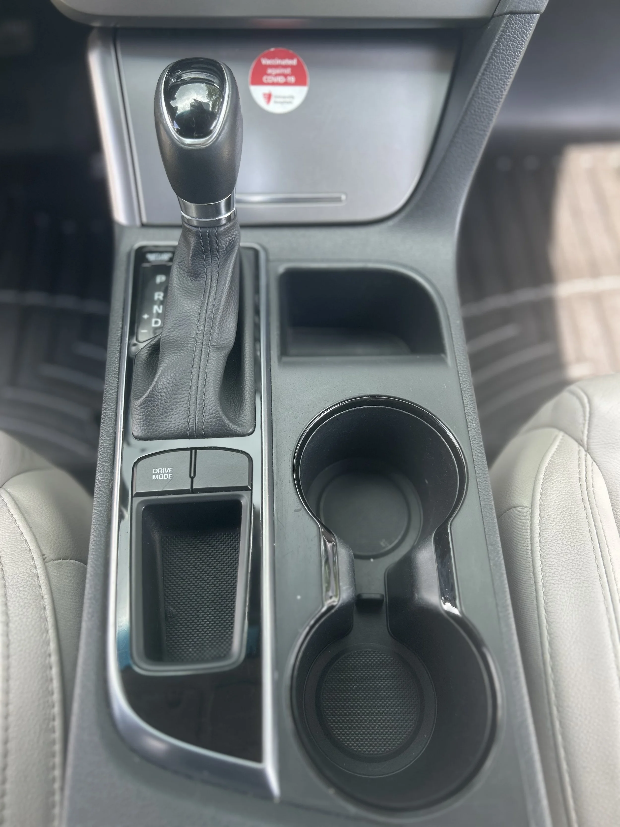 Center console of a car showing gear shift, cup holder, and various compartments, with beige seats on sides.