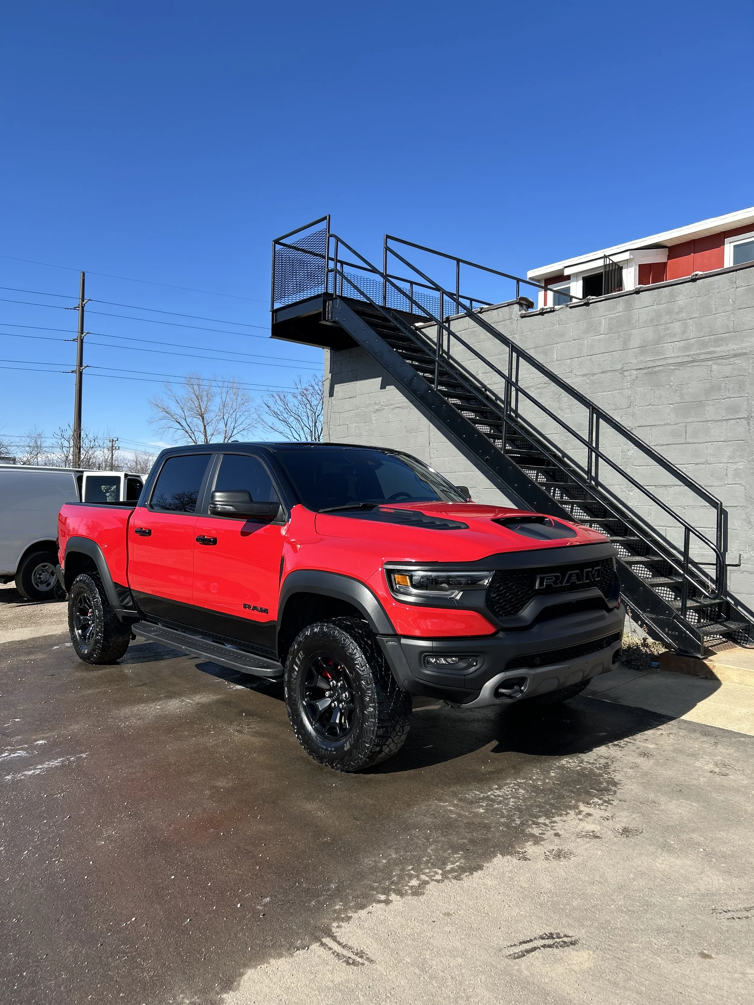 A red and black RAM pickup truck parked on a wet parking lot, with a gray brick building and a black metal exterior staircase leading up to a higher level in the background. Clear blue sky and power lines are also visible.