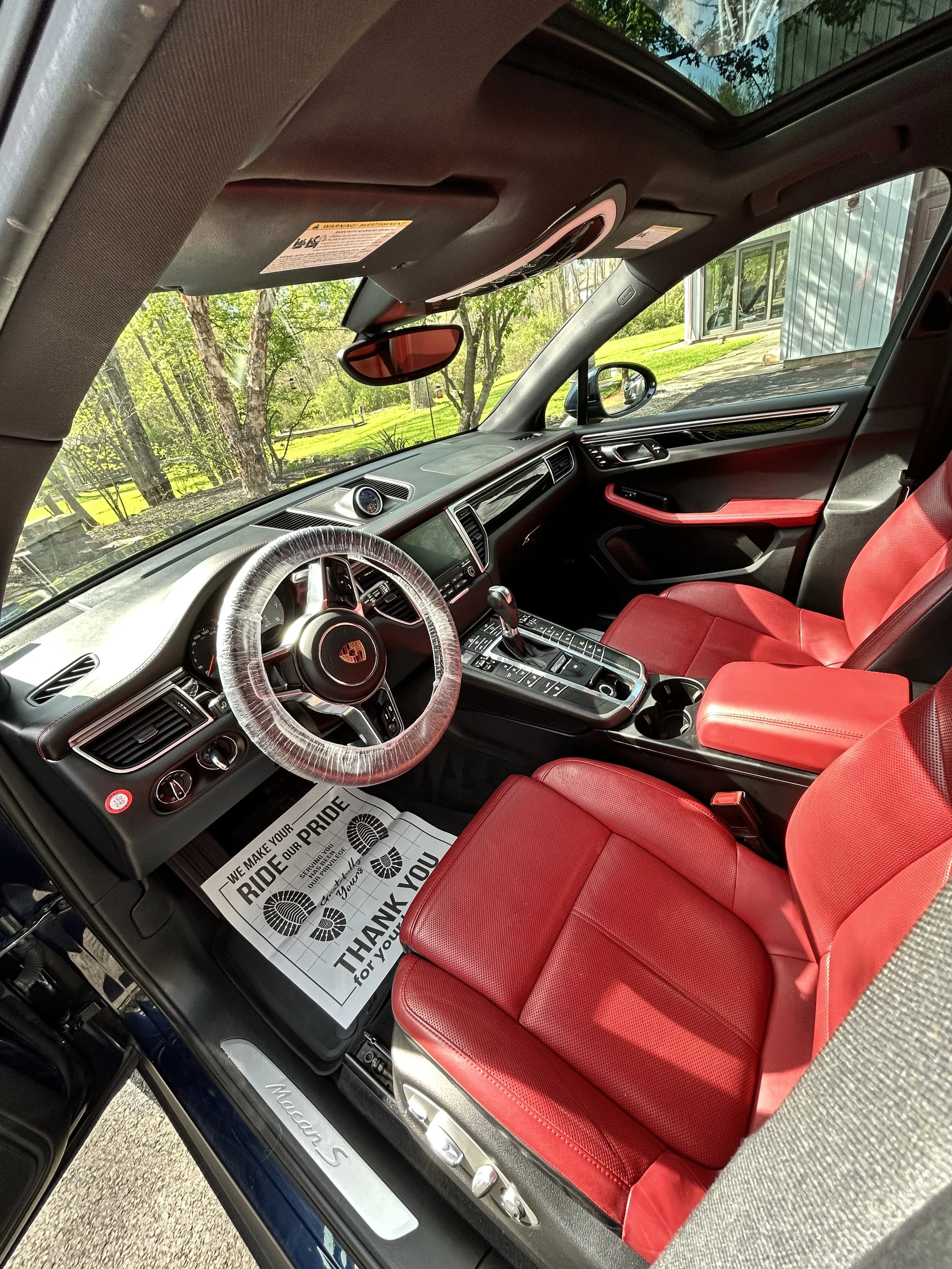 The interior of a Porsche Macan S with red leather seats, black dashboard, and a view of the outside trees and house through the front windshield.