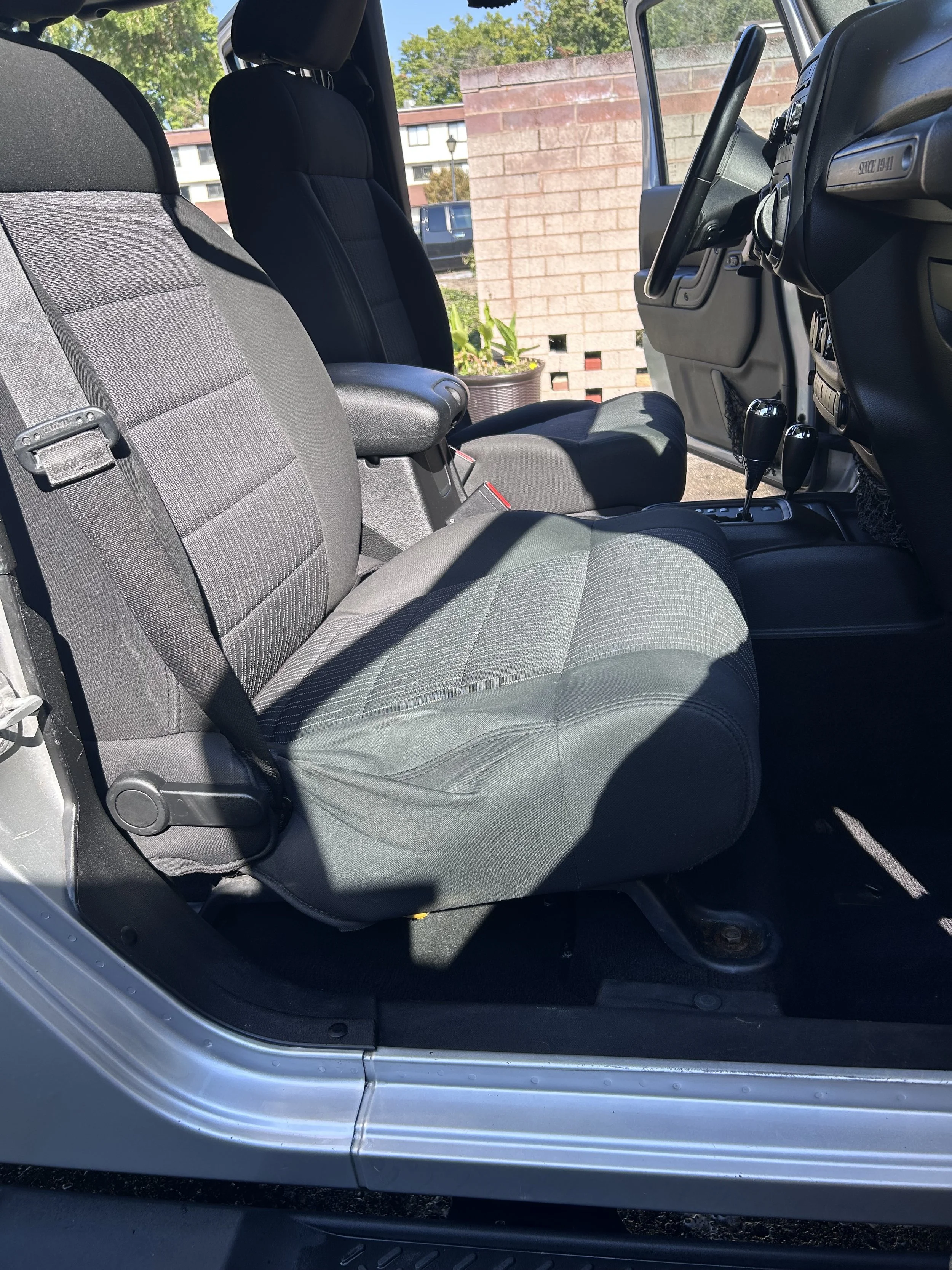 The interior of a vehicle showing the front passenger and driver seats, dashboard, and gear shift. The seats are upholstered in black and gray fabric. The dashboard is black with various controls and vents, and two gear shift levers are visible. The 