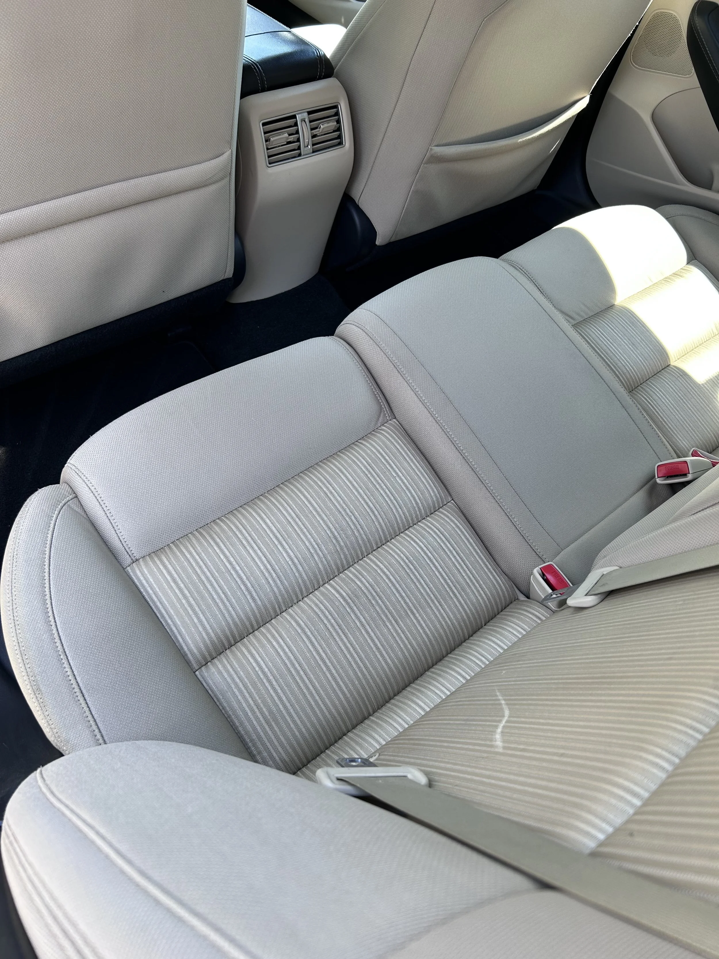 The back seat area of a car with beige and light gray fabric seats, a rear air vent in the center, and visible seatbelt buckles.