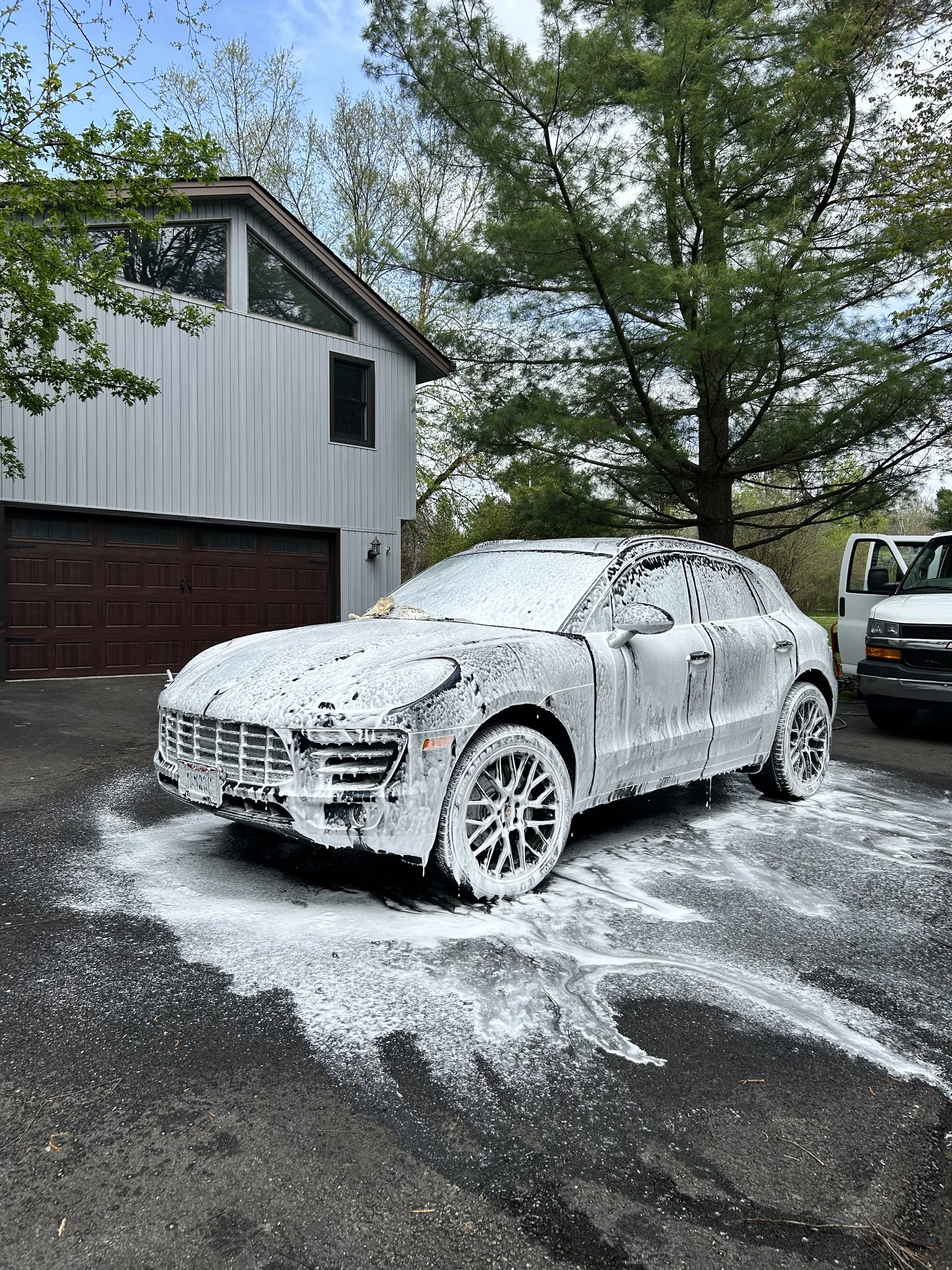 A silver car covered in white foam soap parked on a driveway. A large tree and a house with a garage are visible in the background.