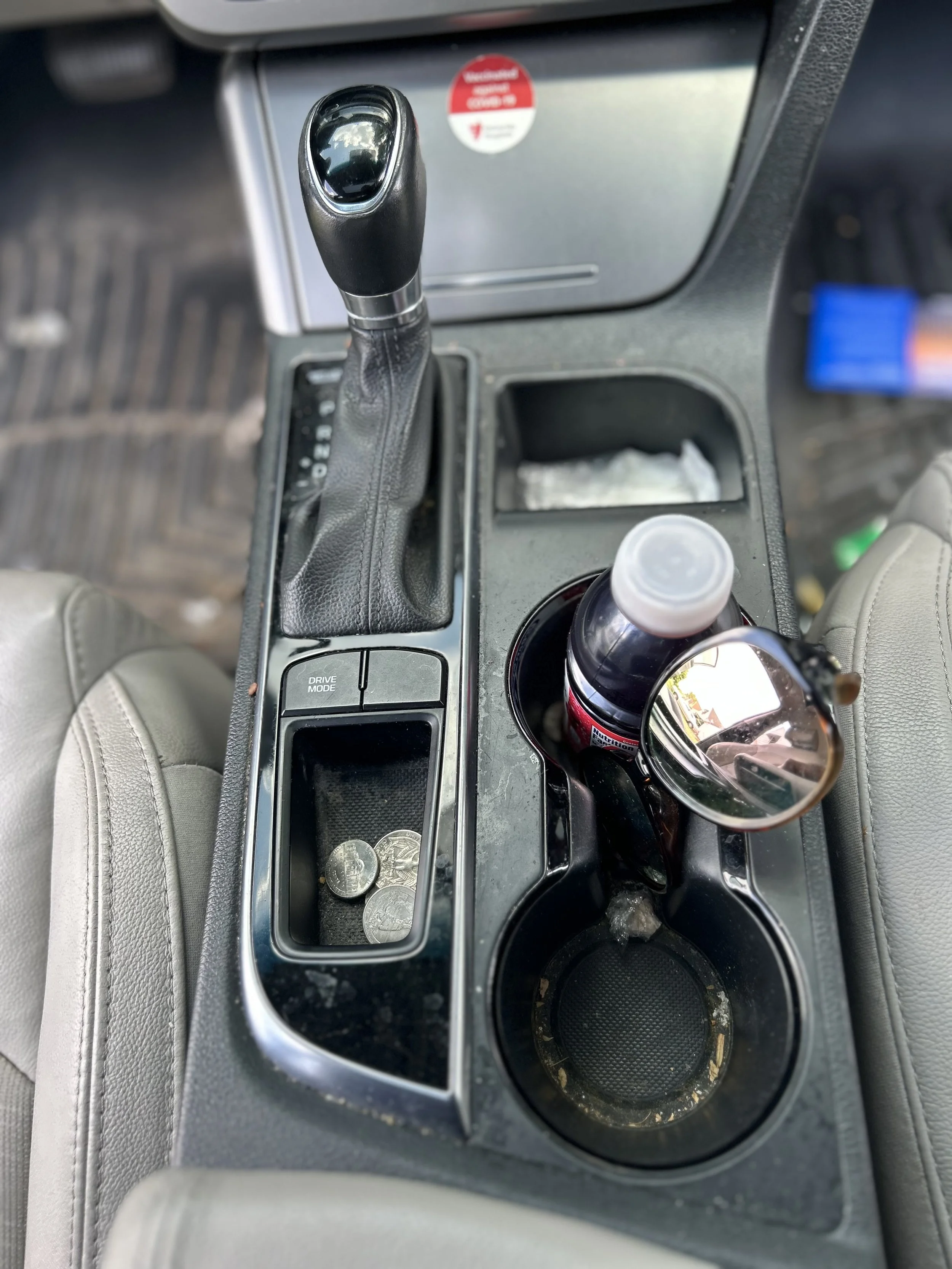 Center console of a car with a gear shift, some coins, a beverage bottle, sunglasses, and empty cup holders.
