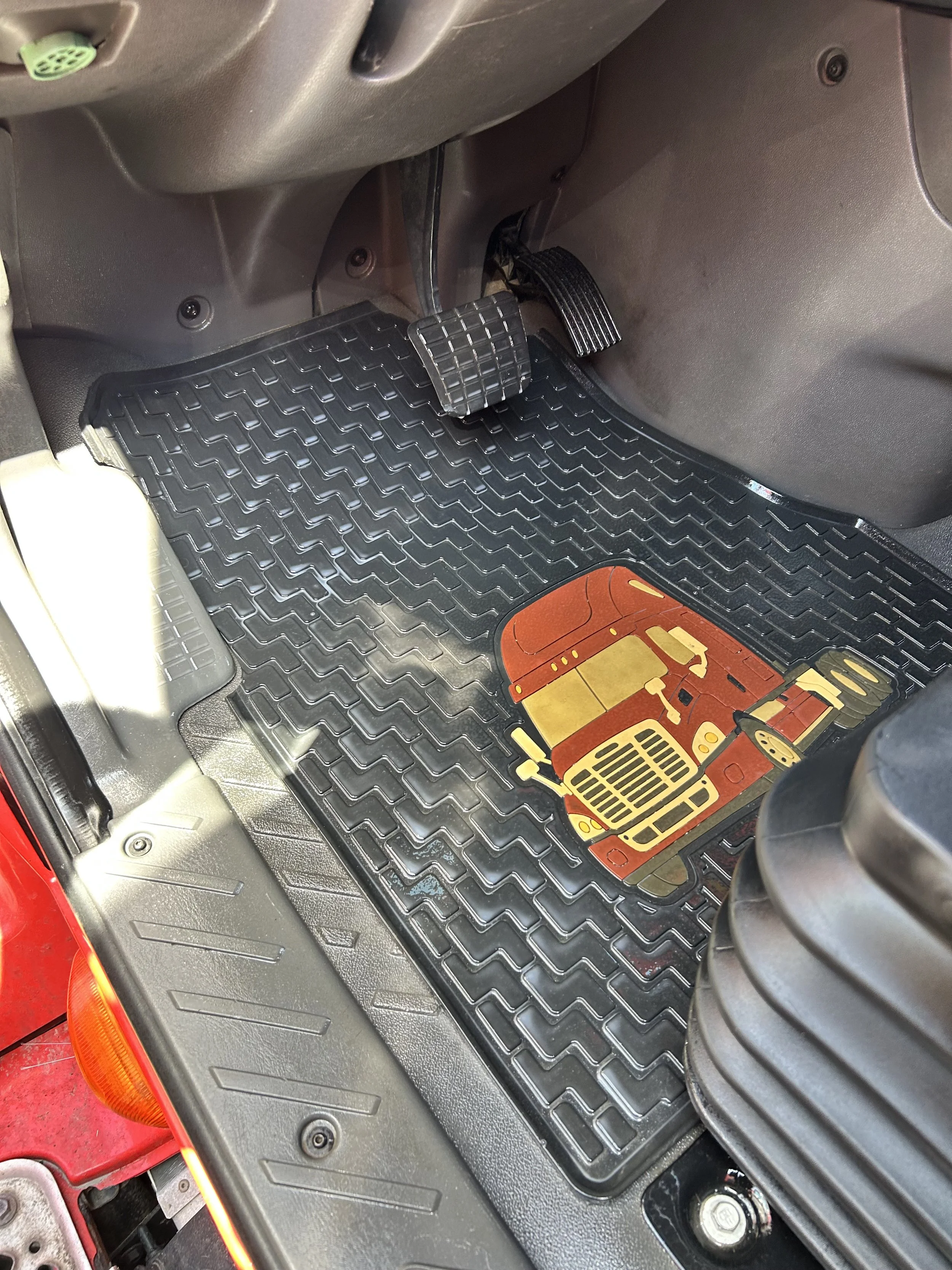 Interior of a vehicle showing the brake pedal, gas pedal, and a black rubber floor mat with a vehicle-themed design.