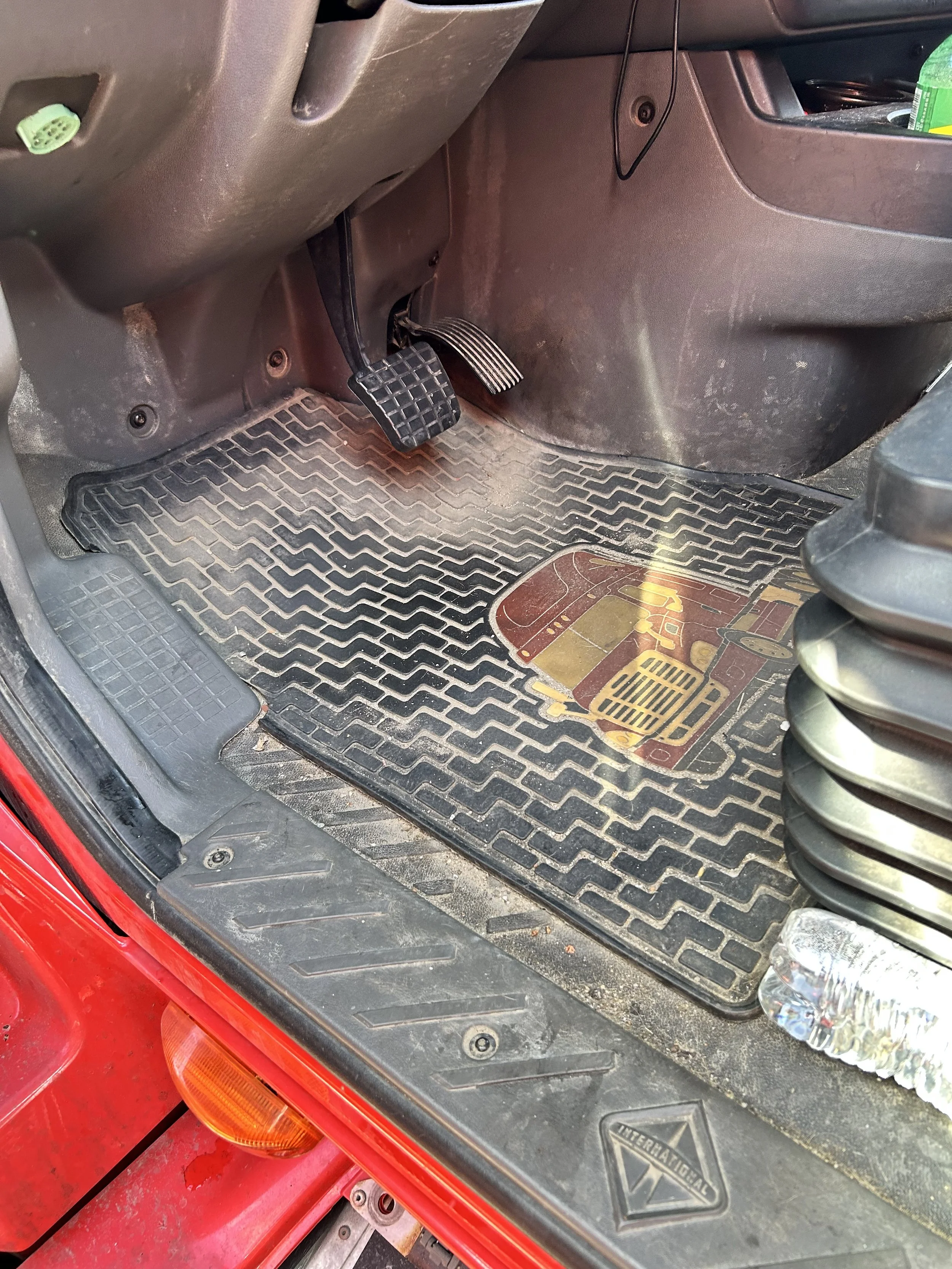 The image shows the driver's side footwell of a vehicle, with visible brake and accelerator pedals, a floor mat, and part of the car's interior including a plastic water bottle and a container in the storage compartment. The floor and pedals appear s