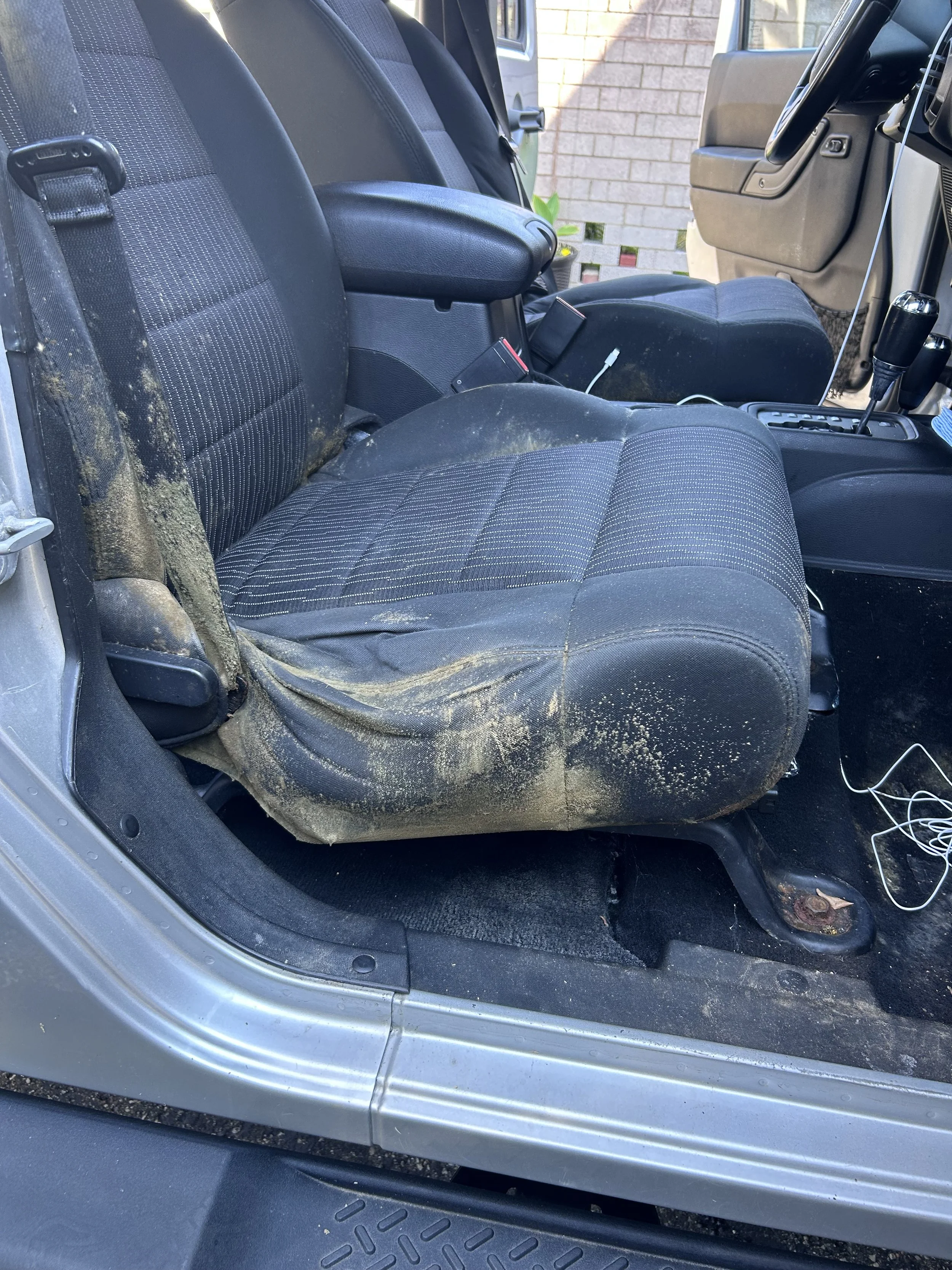 The front passenger seat of a vehicle with dirt and mud stains, showing some dirt on the seat and floor area, with a brick wall visible outside the vehicle.