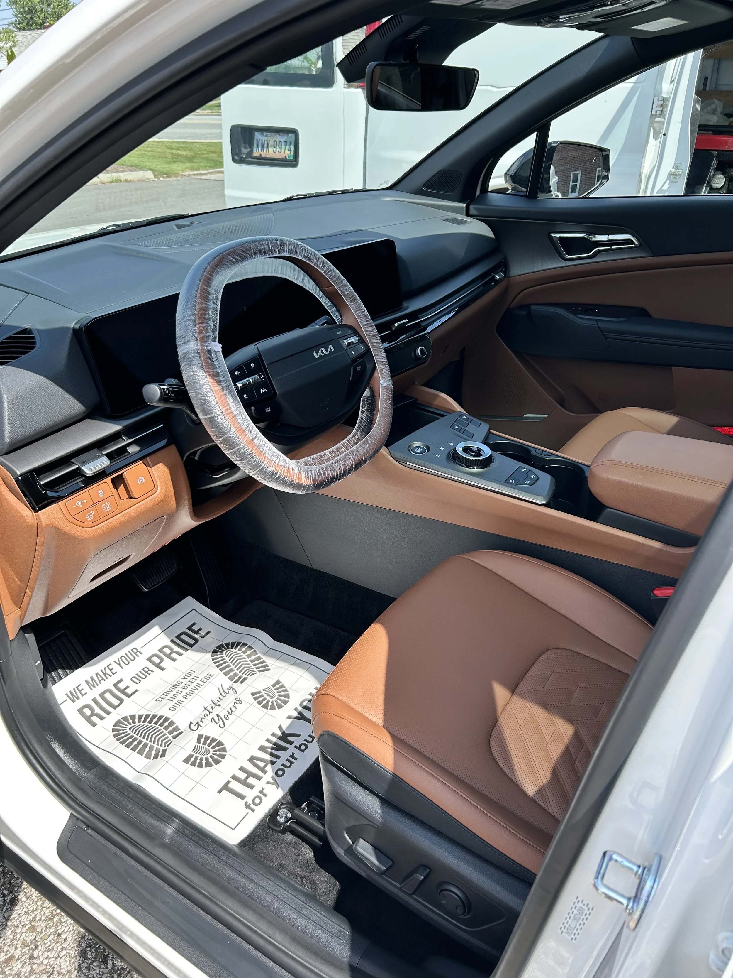 Interior of a modern vehicle showing the dashboard, steering wheel with plastic cover, and tan leather seats. The door is open, revealing part of the exterior environment with a white truck and some greenery visible outside.