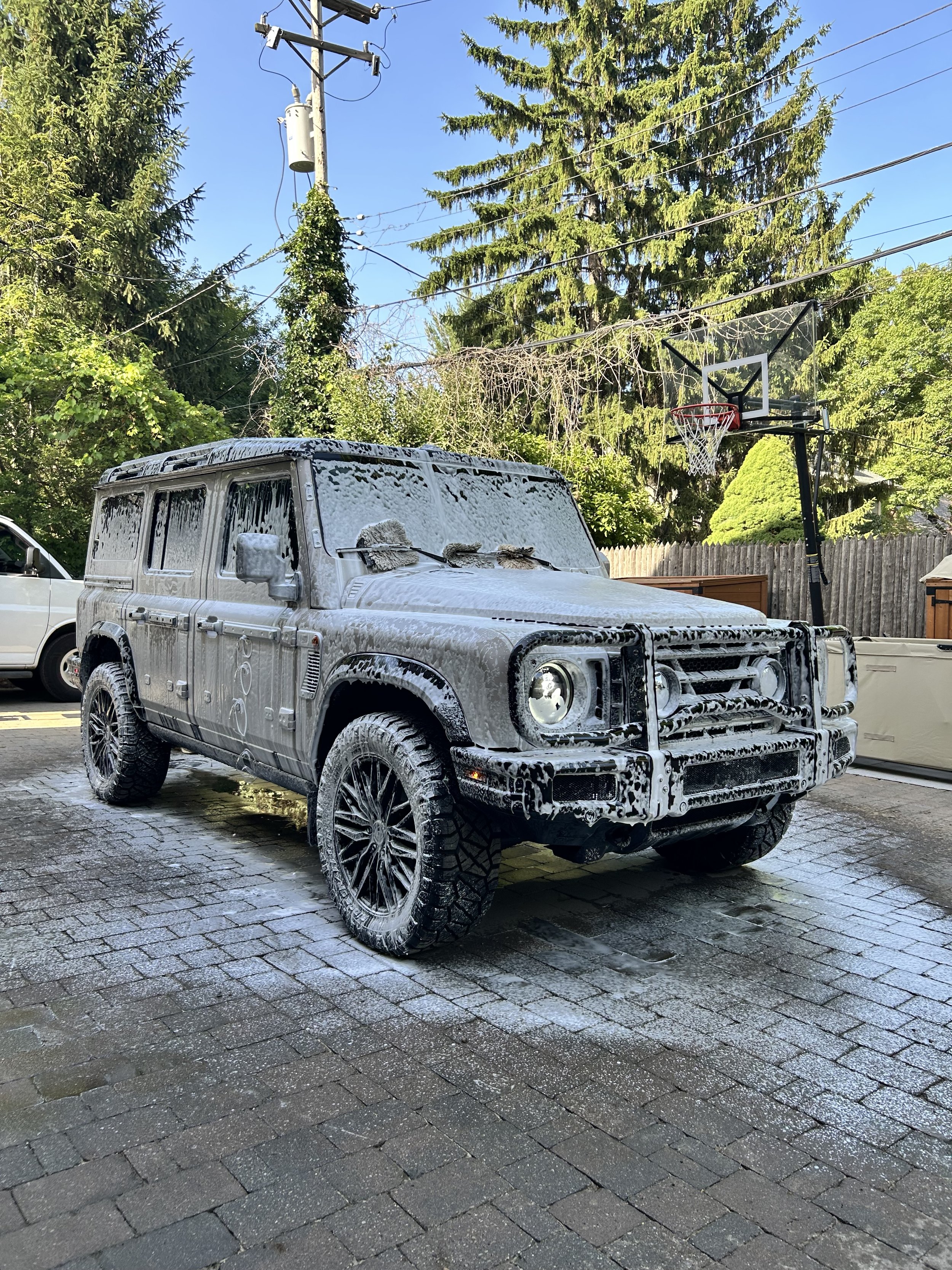 A black SUV covered in soap and water during a car wash, with soap suds on the hood, windshield, and roof. The vehicle is parked on a wet, cobblestone driveway, and behind it are trees, a wooden fence, and a basketball hoop.