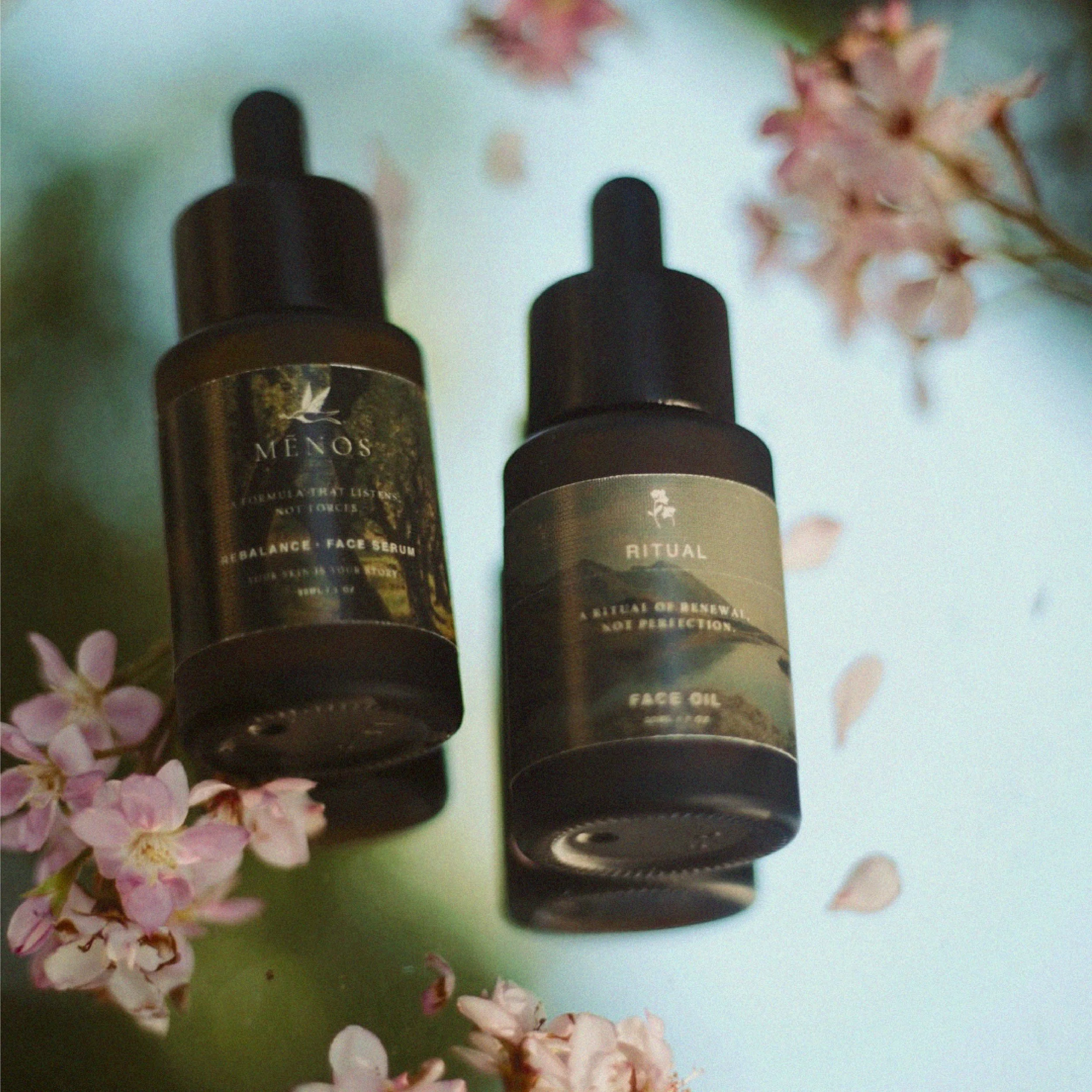 Two MĒNOS dropper bottles with sage green labels lying among pink petals