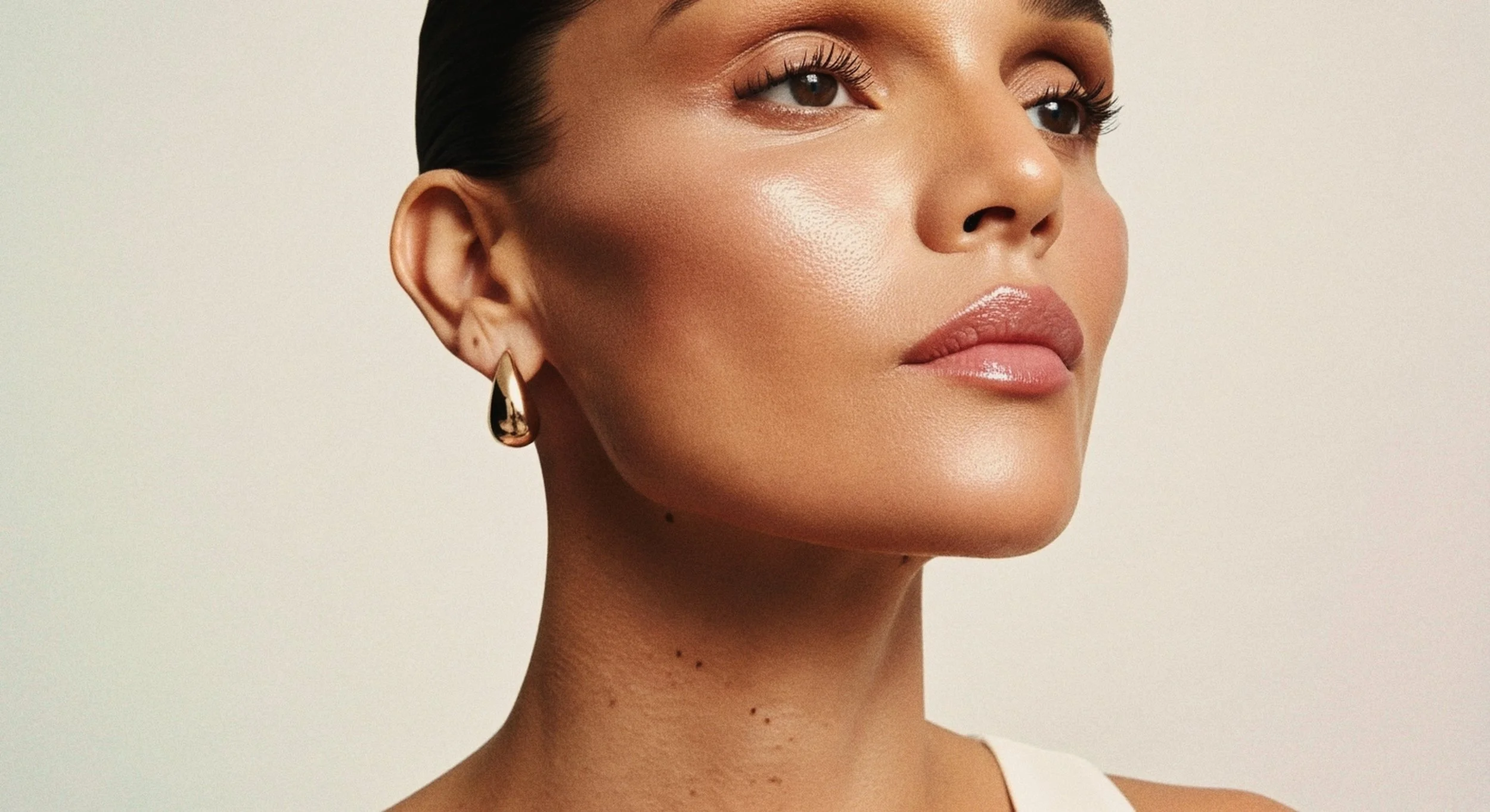 Close-up of a woman's face with smooth skin, wearing gold teardrop-shaped earrings, with neutral makeup and subtle pink lips, against a plain background.