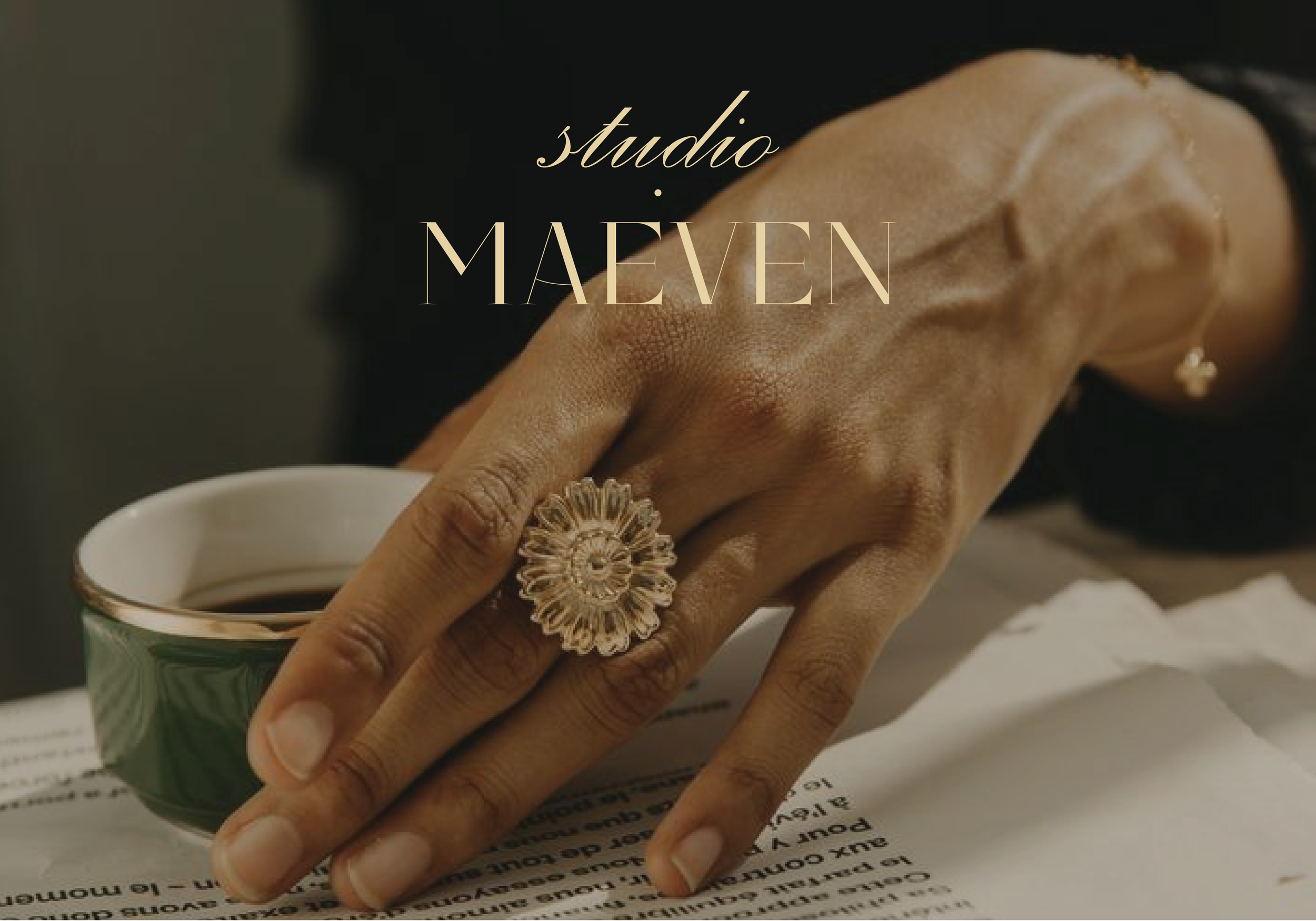 A hand with a gold ring and earring holding a coffee cup, with a piece of paper underneath, and the words 'studio MAEEN' overlaid on the image.